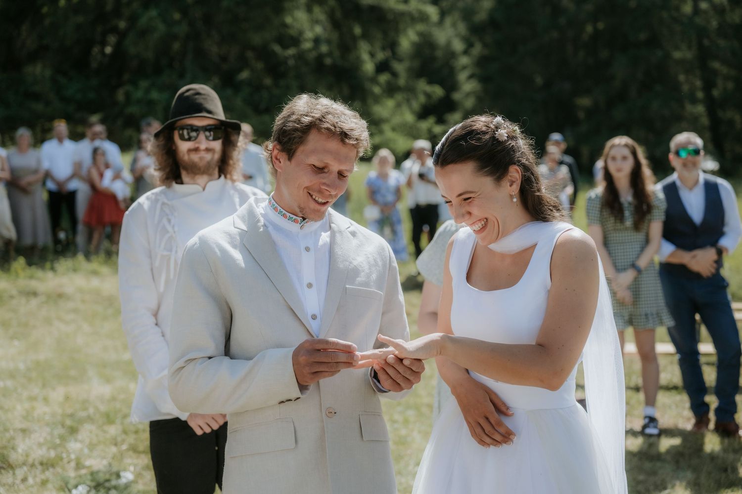 Bride and groom exchanging rings outdoors at a sunny garden wedding ceremony with guests watching in background.