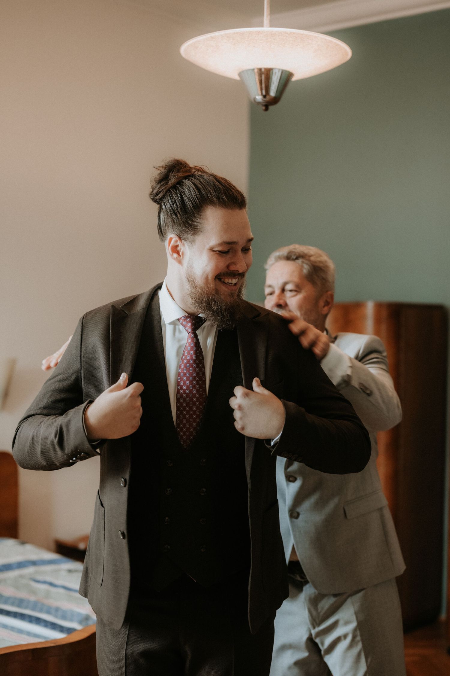 Groom in dark suit adjusting jacket lapels while smiling, assisted by man in gray suit in elegant room.