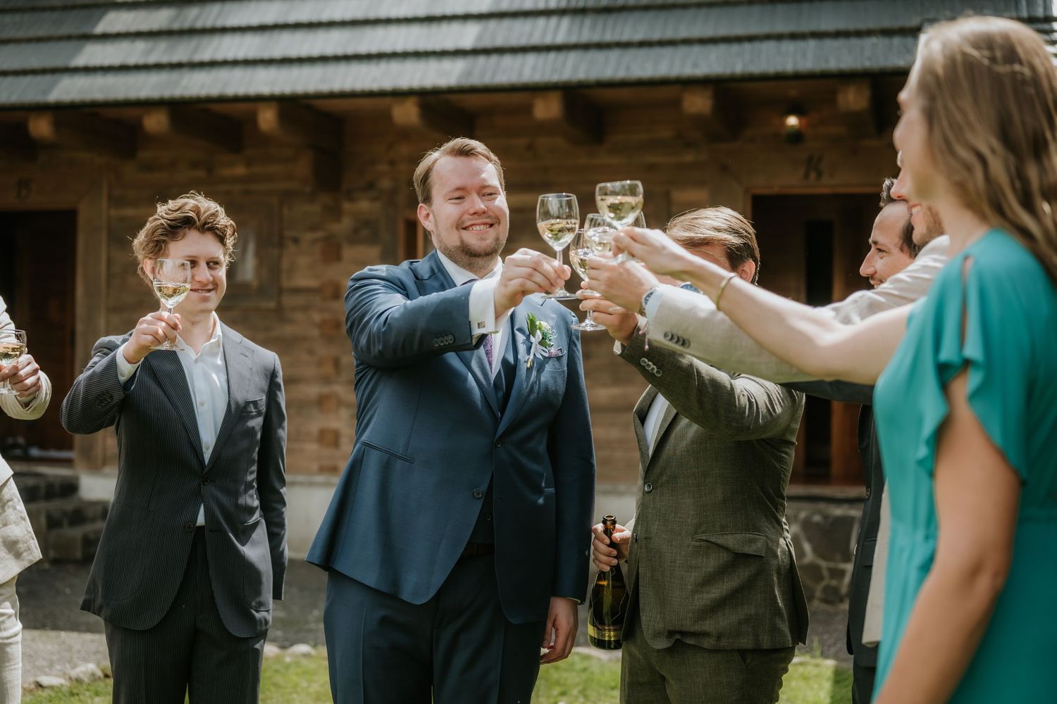 Wedding guests raising champagne glasses in a joyful outdoor toast near a rustic stone building.