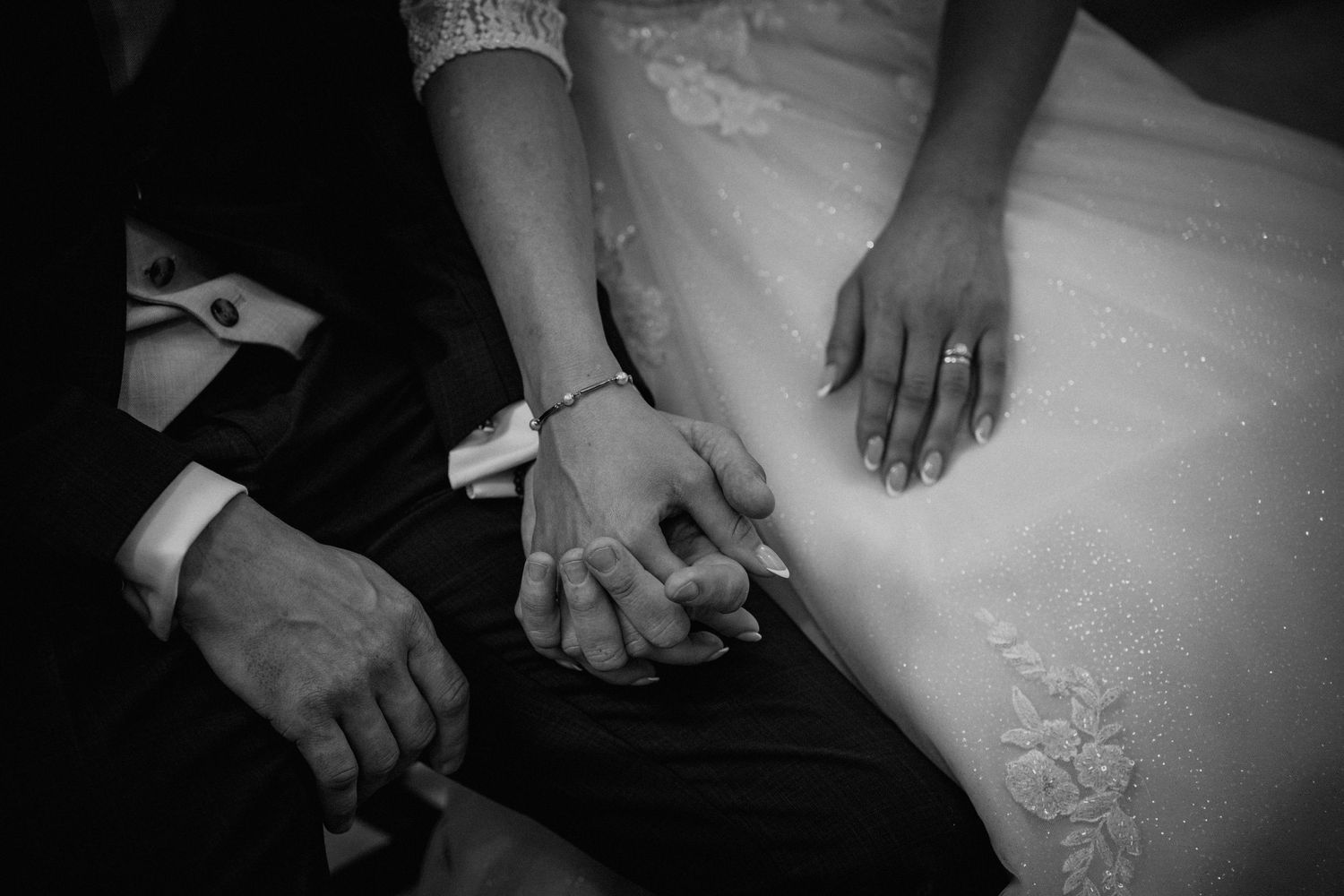 Black and white photo of newlyweds holding hands, wedding rings visible, bride in white lace gown beside groom in dark suit.