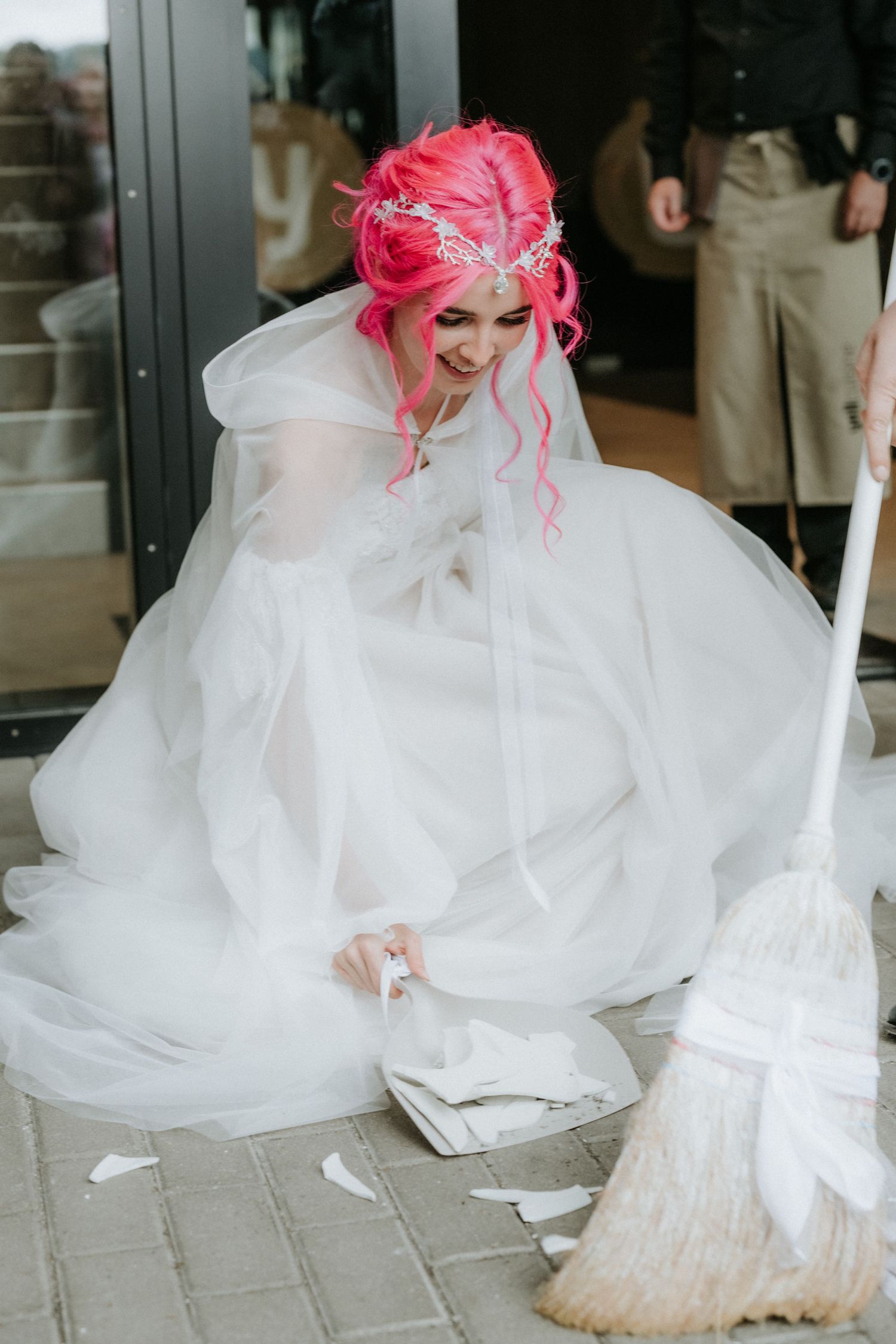 Bride with pink hair in white gown sweeping broken pieces on pavement with a broom outdoors.
