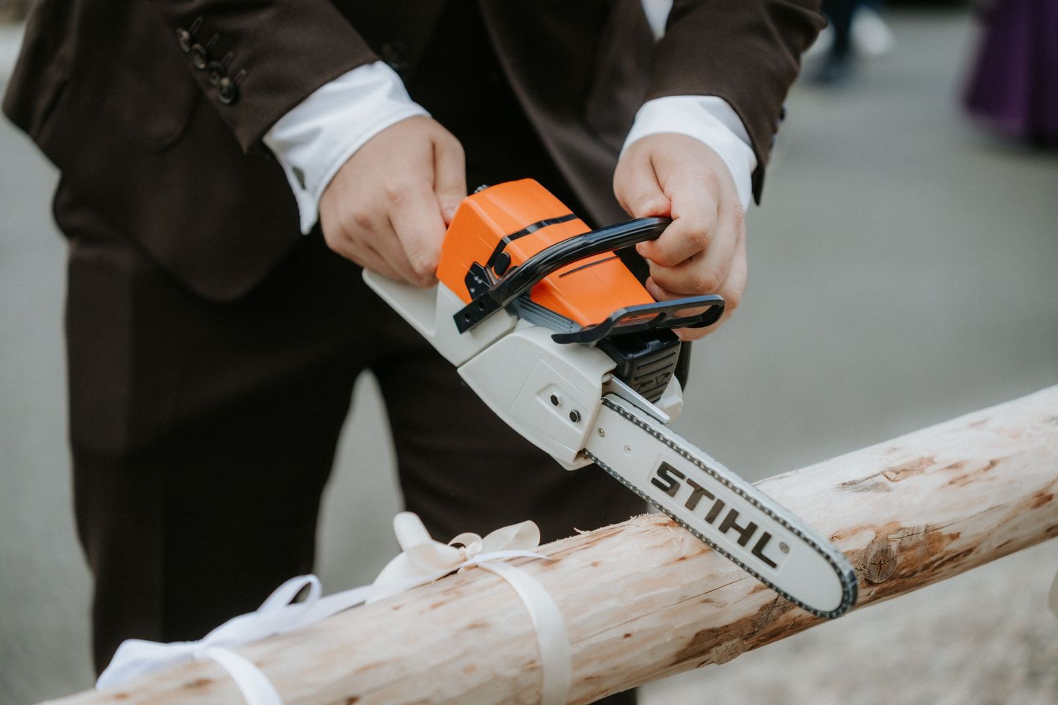 Man in suit using a Stihl chainsaw to cut a log outdoors at a formal event.