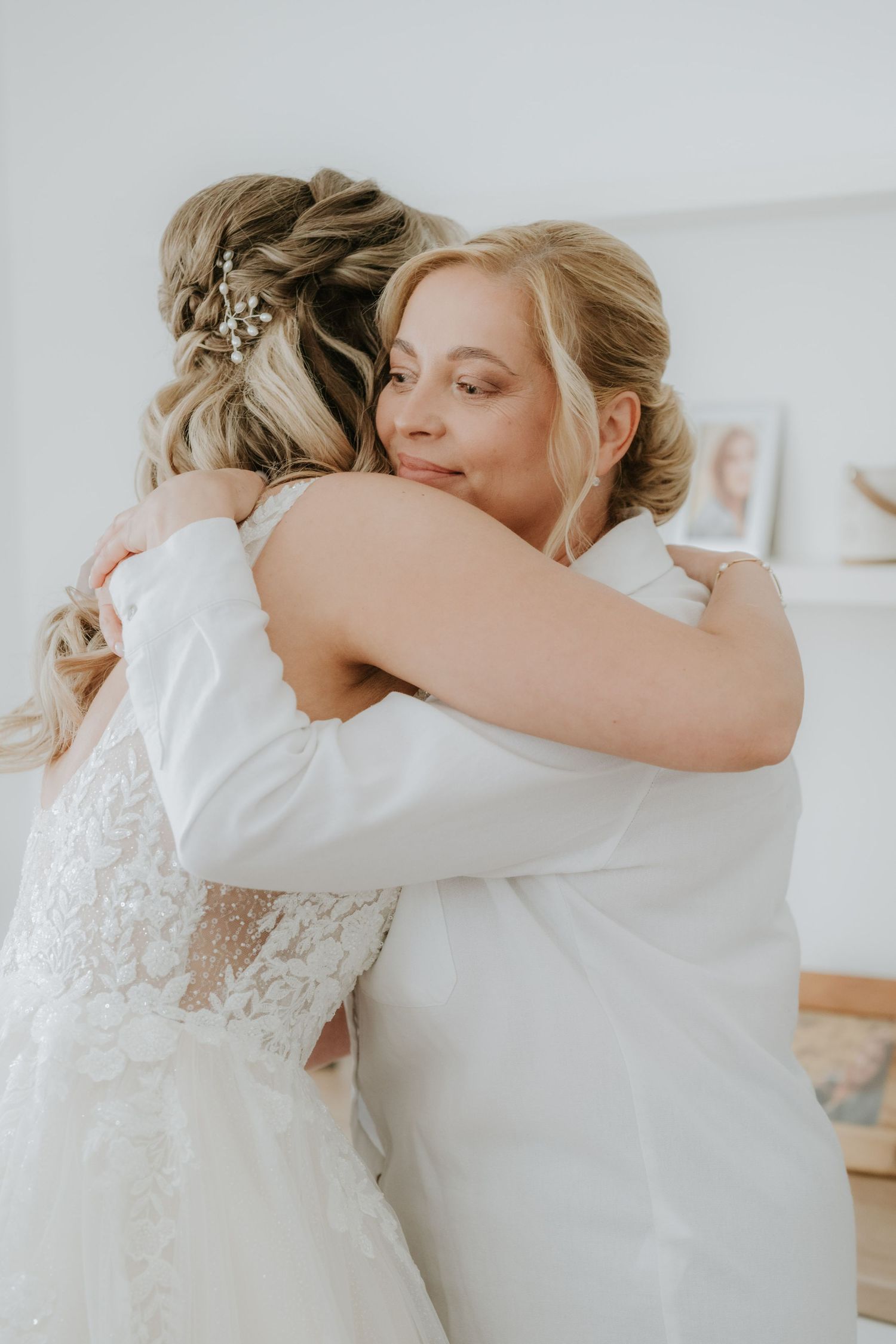 Two blonde women in white dresses share an emotional hug during a wedding day preparation moment.