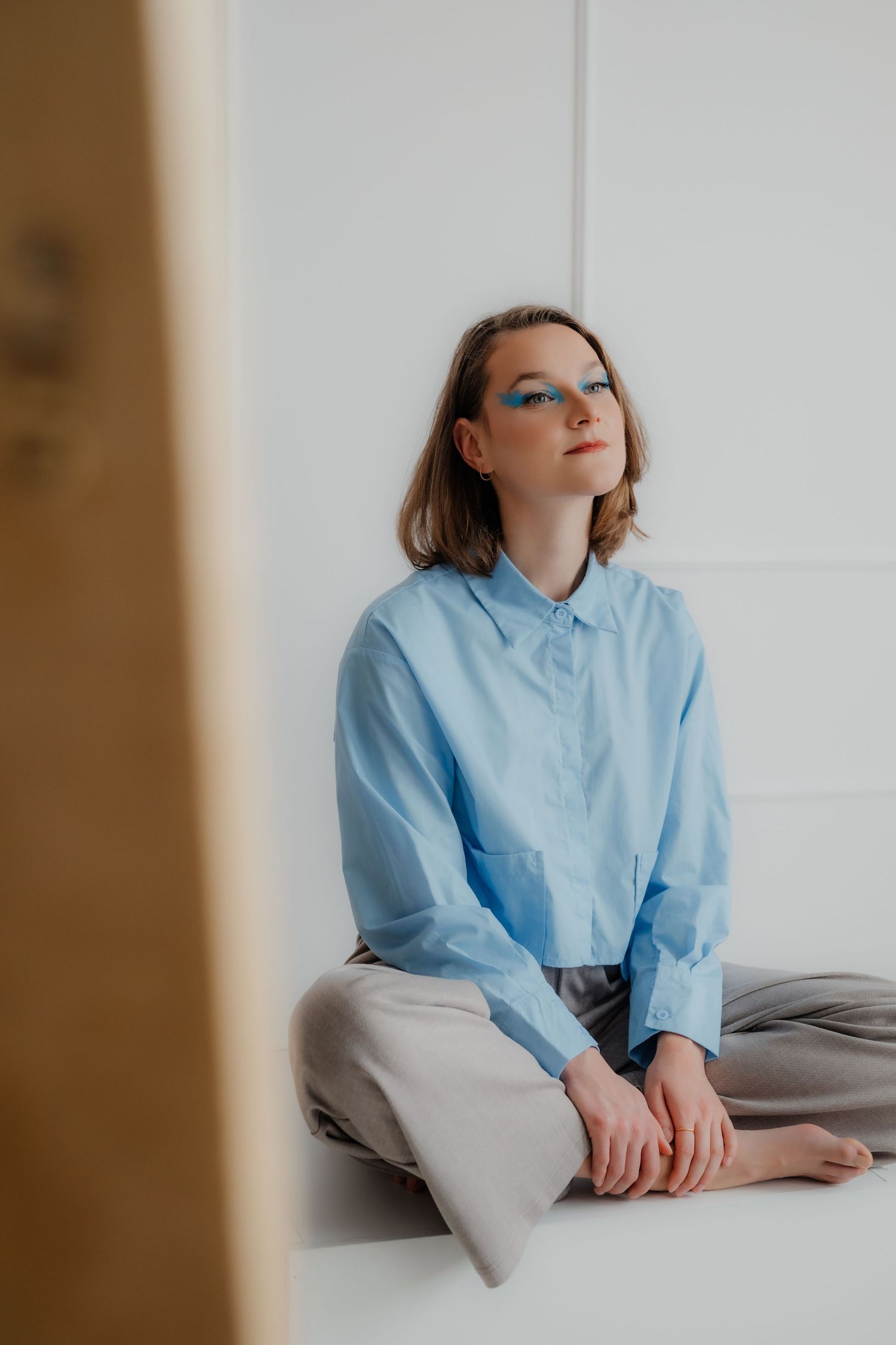 Young woman with blue graphic eye makeup sitting cross-legged on white surface in light blue shirt and gray pants.