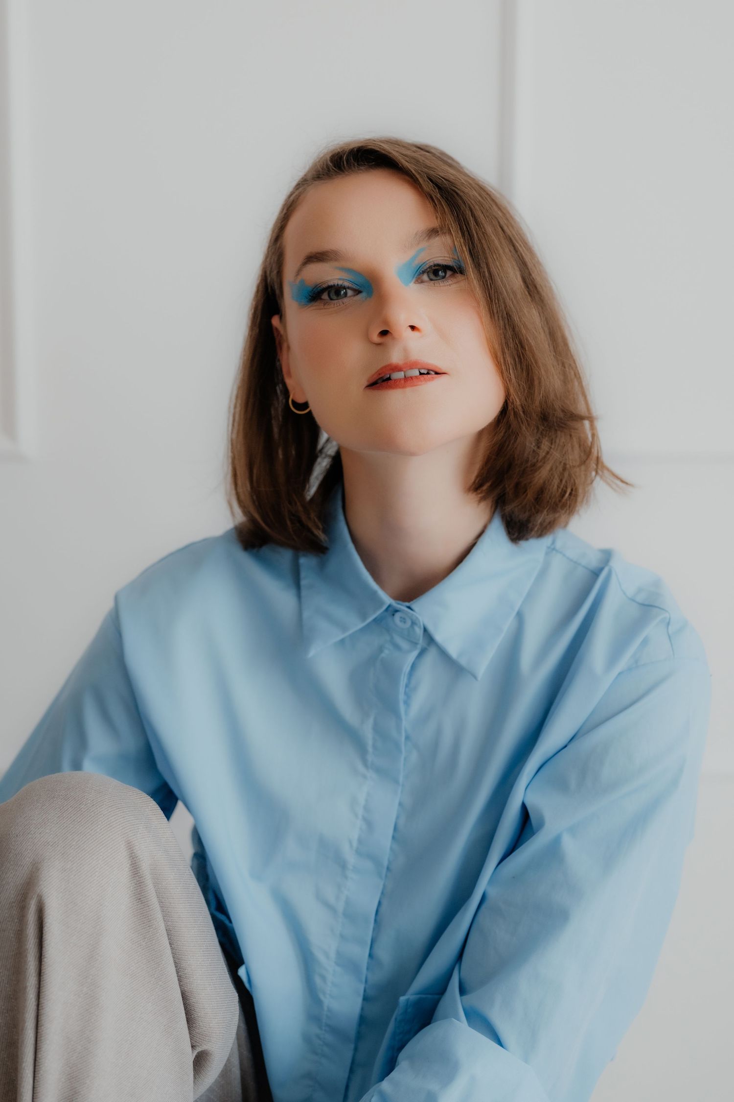 Young woman with bold blue eye makeup wearing light blue oversized shirt sitting against white wall.