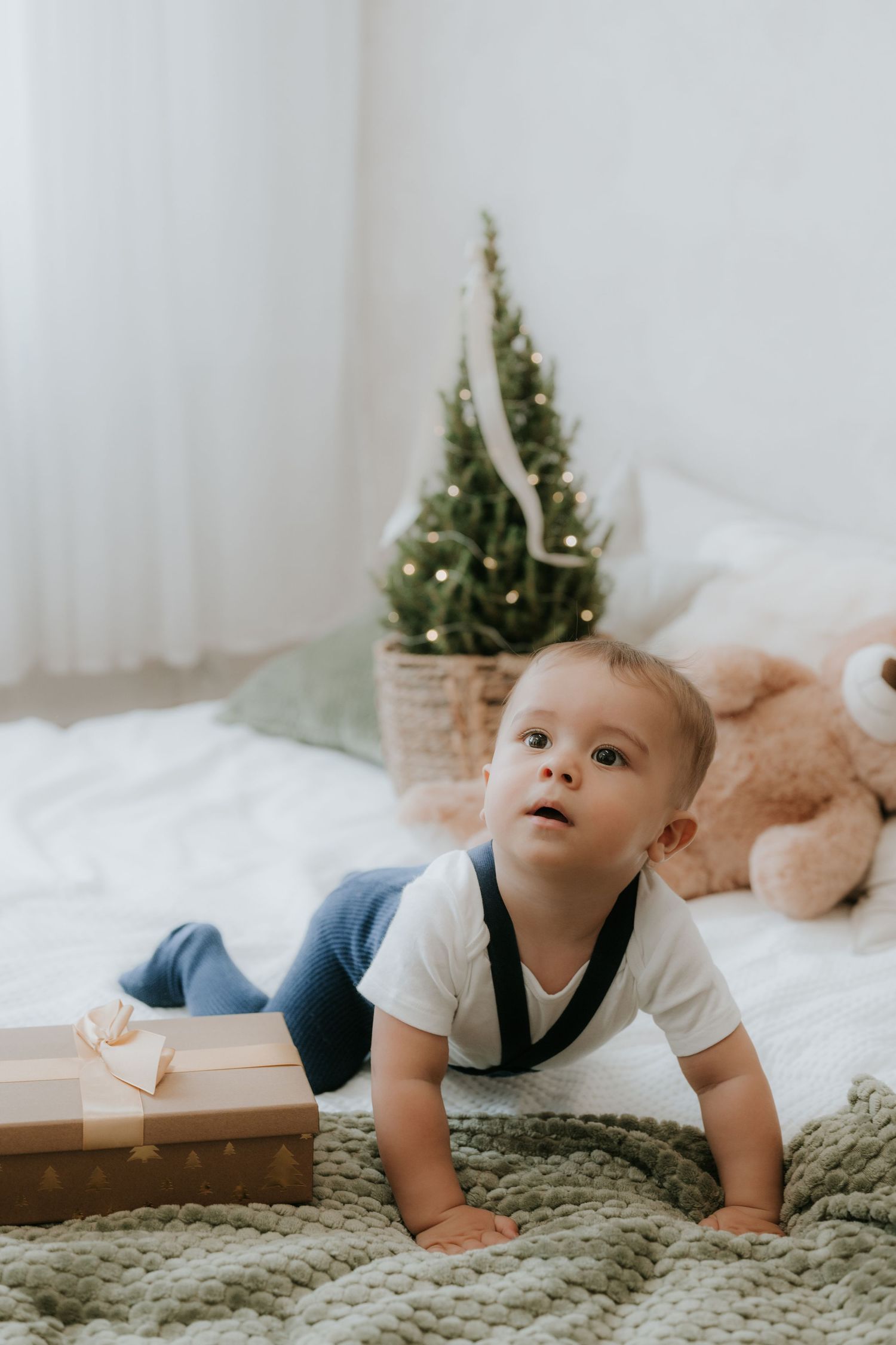 Curious baby in white bodysuit crawling on bed near gift box and Christmas tree with teddy bear.