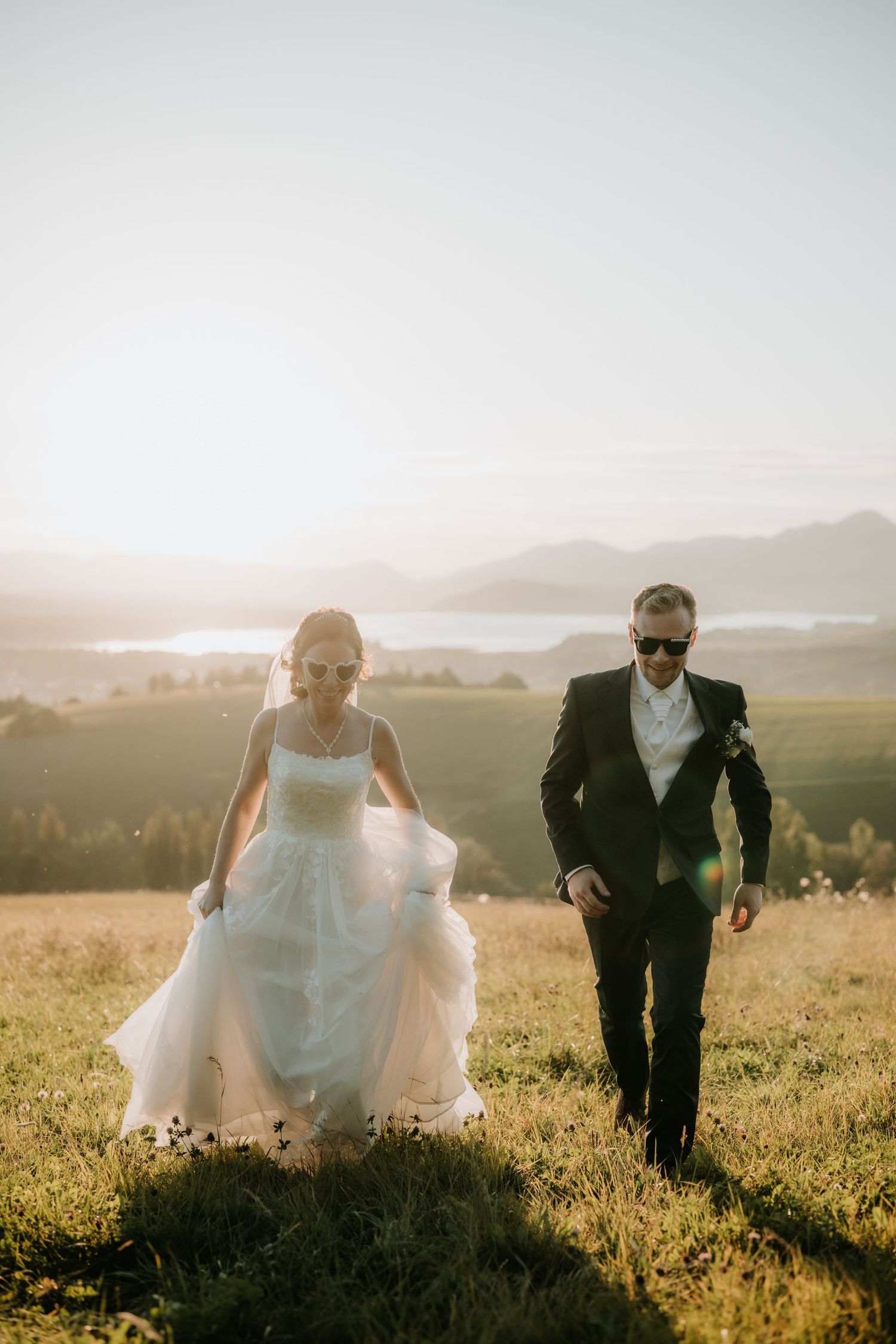 Bride in white gown and groom in black suit walk through sunlit meadow with mountain lake backdrop.