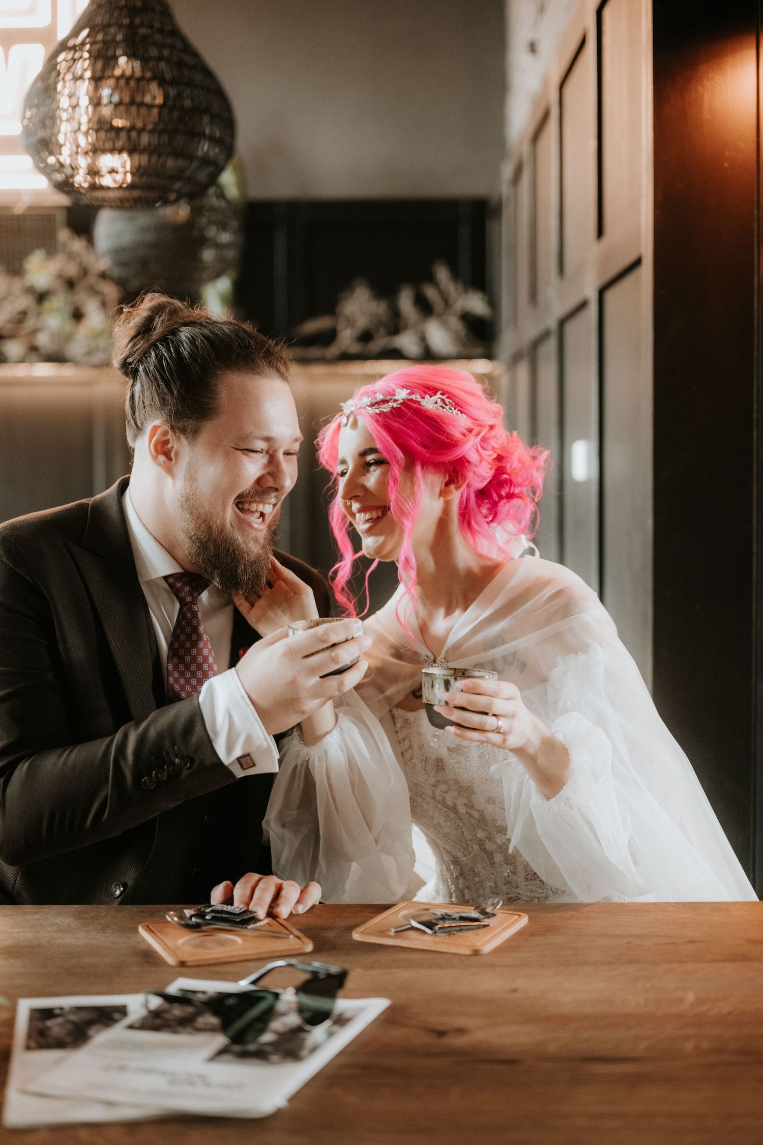 Joyful newlyweds sharing a laugh over espresso shots at a wooden table in a cozy venue.