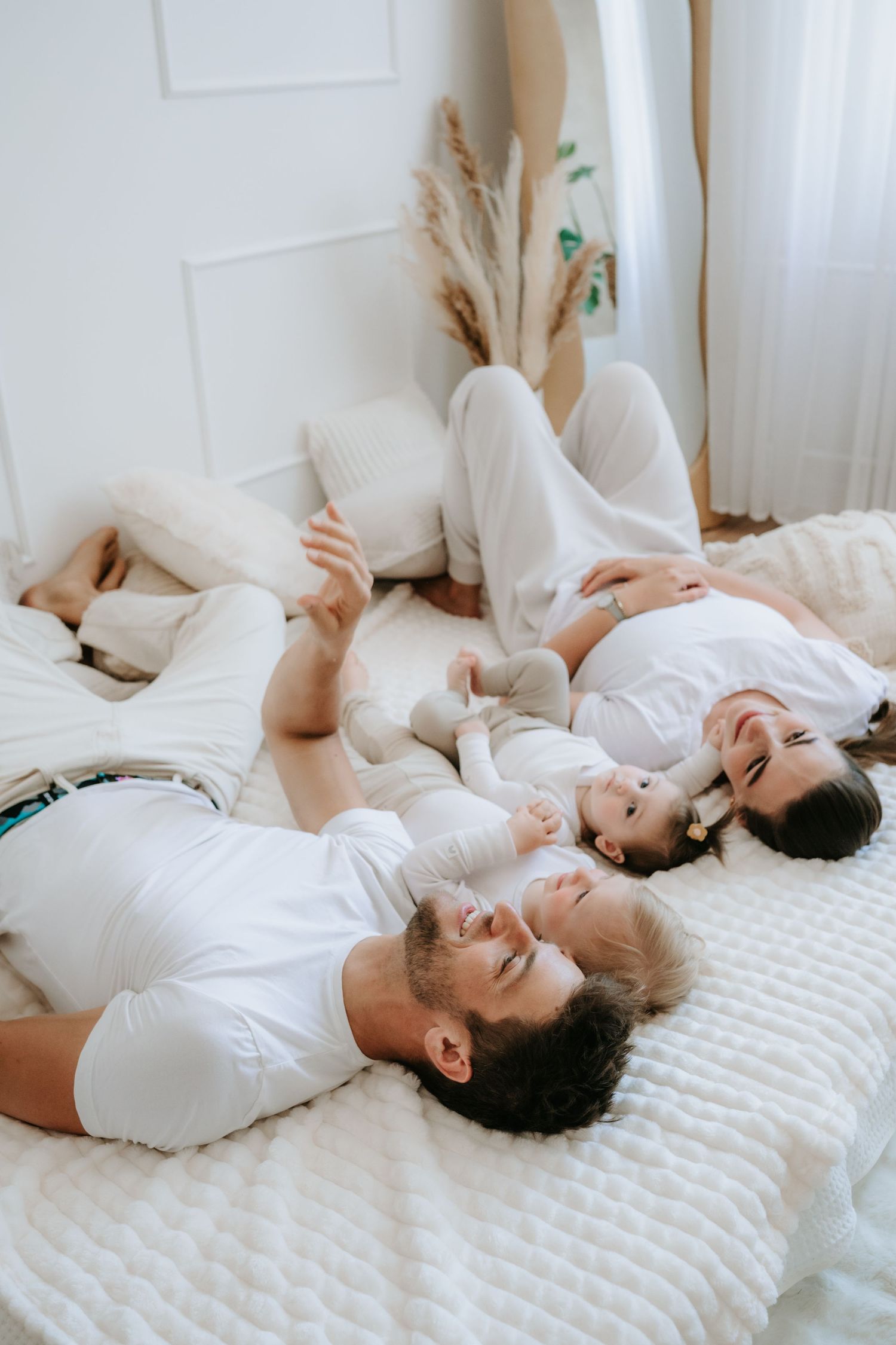 Young family of four lying together on white bed laughing in bright minimalist bedroom.