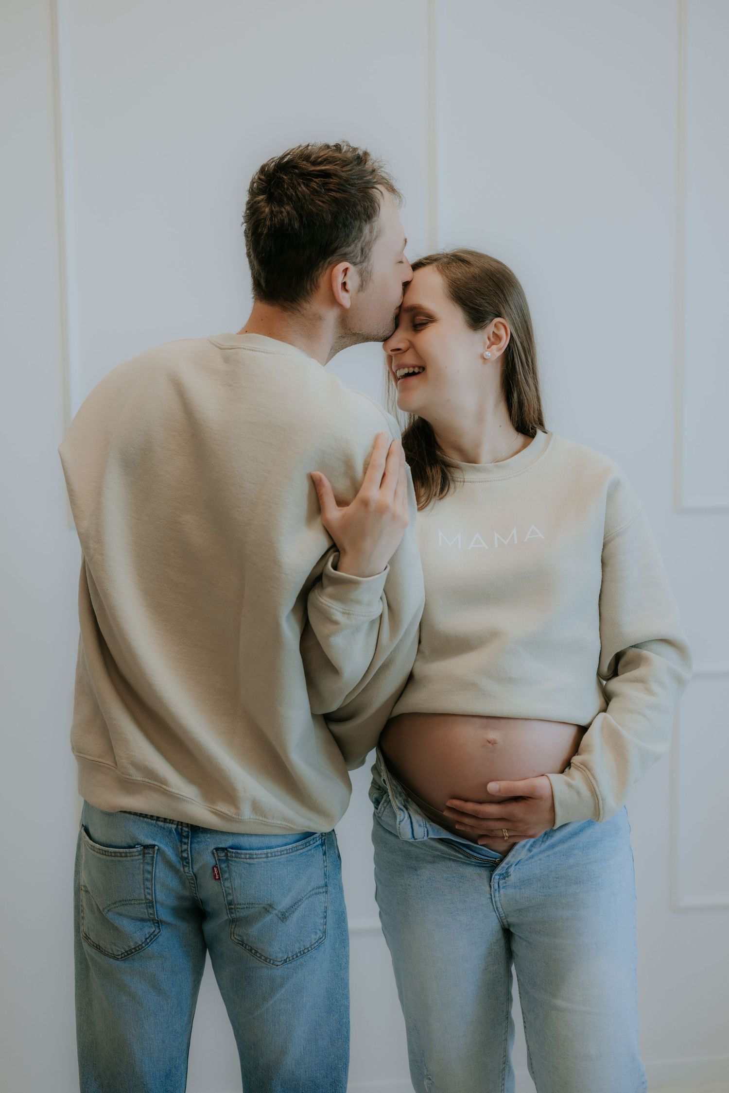 Pregnant woman in MAMA sweatshirt smiles as partner kisses her cheek, both in jeans.