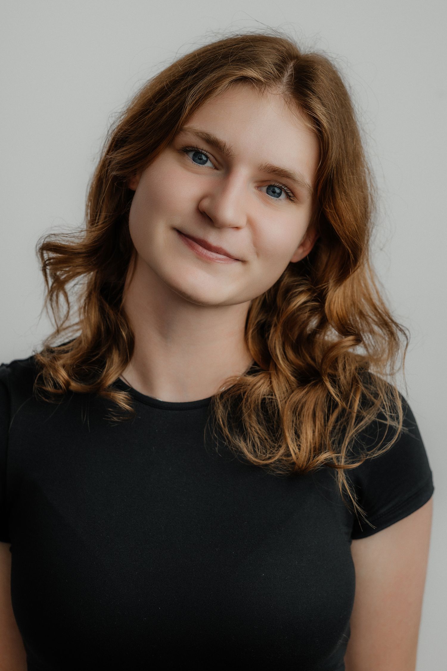 Young woman with wavy auburn hair and blue eyes smiling softly, wearing a black t-shirt against a gray background.