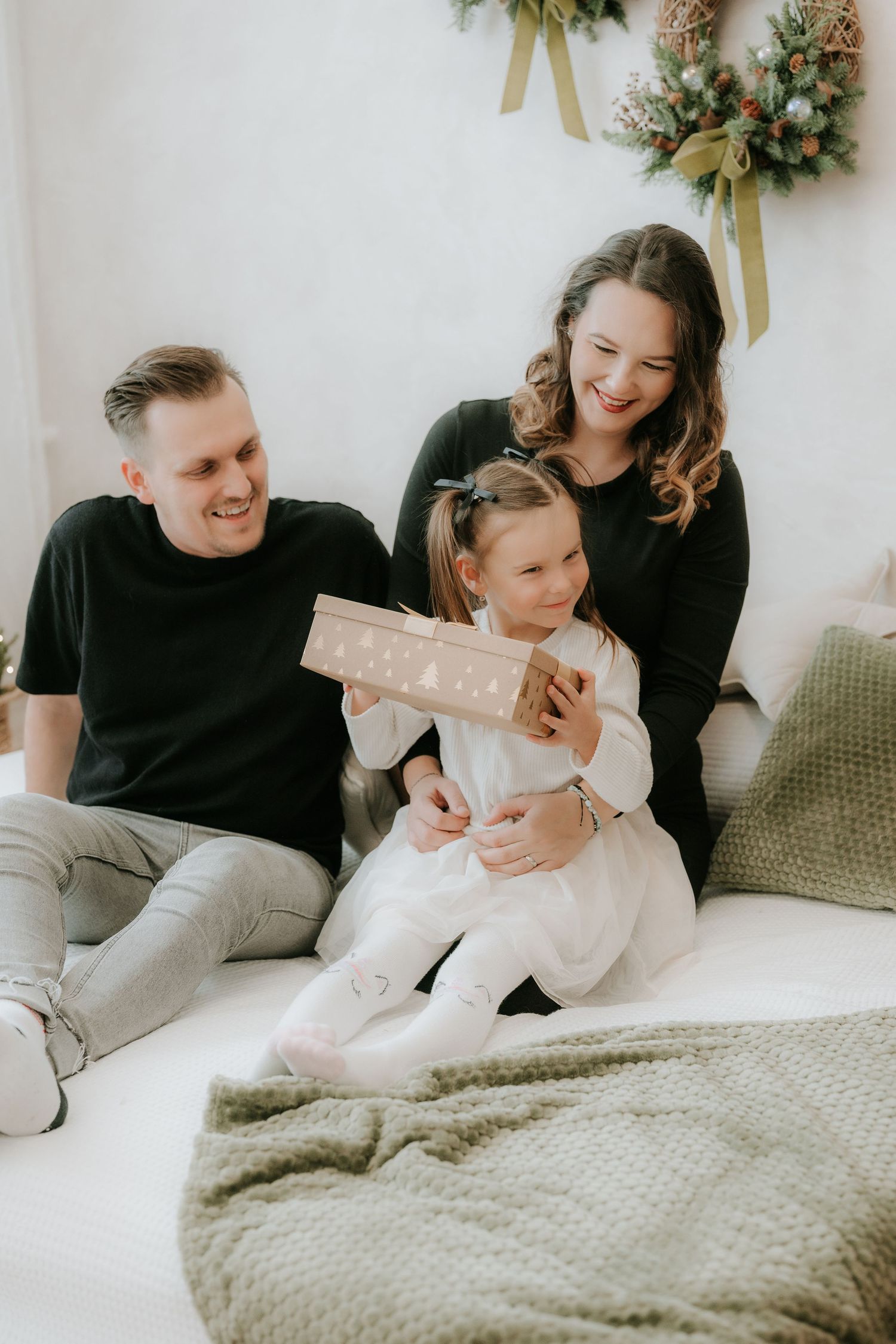 Joyful family with little girl opening Christmas gift on white bed with festive decorations.