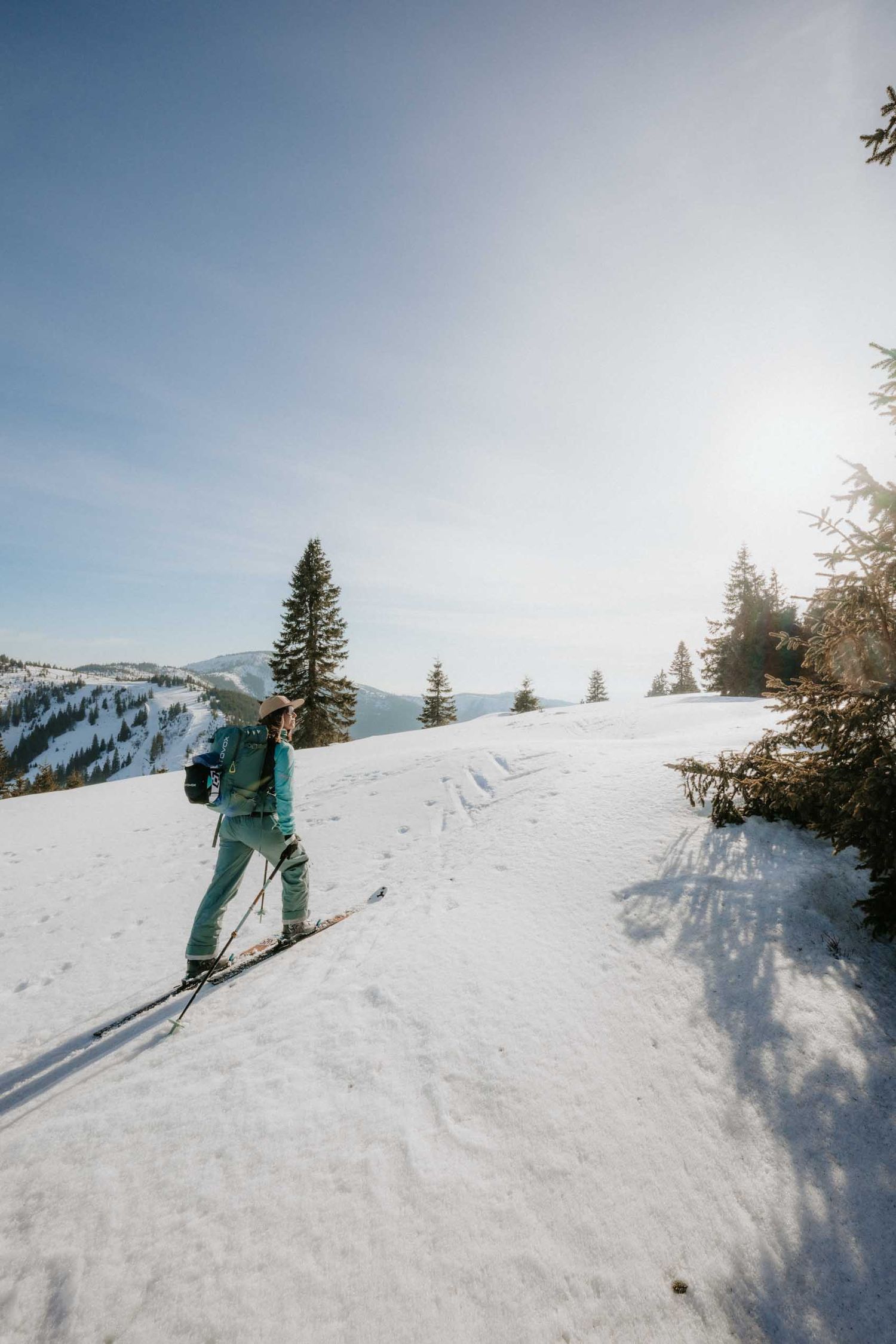 Female backcountry skier in teal gear ascending sunny snow-covered mountain trail with pine trees.