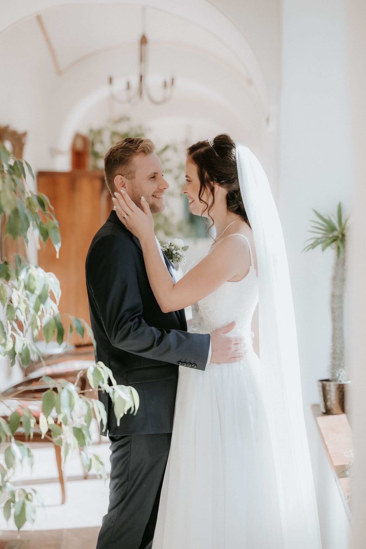 Bride and groom share a tender moment indoors, she in a white gown and veil, he in a black suit, smiling at each other.