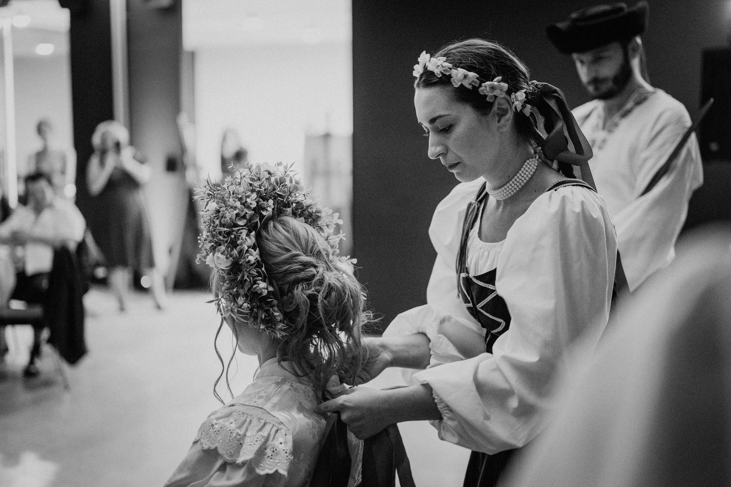 Black and white photo of a woman in folk costume adjusting a flower crown on a young girl at a traditional ceremony.