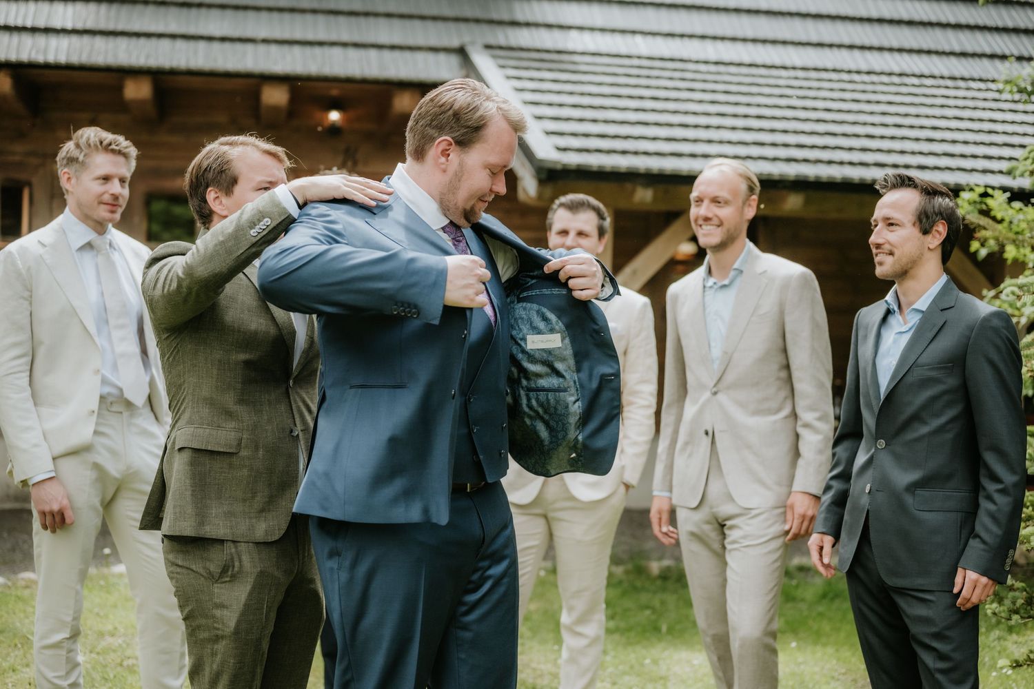 Groomsmen helping groom put on jacket outdoors at wedding venue surrounded by men in suits.