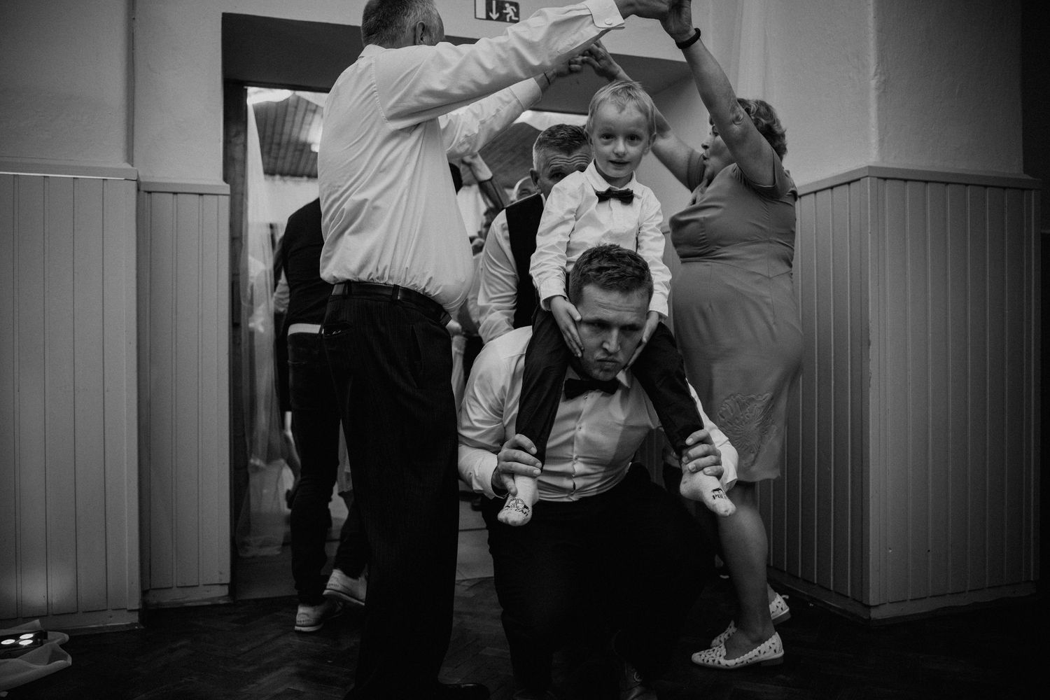 Black and white wedding reception photo of man carrying child on shoulders while guests dance and celebrate.
