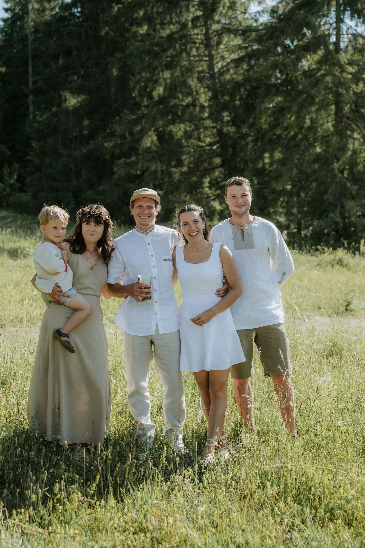A happy family group of five poses in a sunny green meadow surrounded by tall pine trees, dressed in light summer outfits.