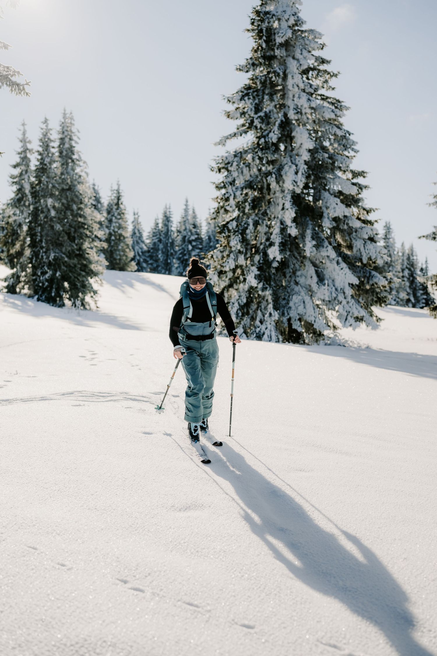 A skier ascends a snowy mountain slope surrounded by snow-covered evergreen trees on a sunny winter day.