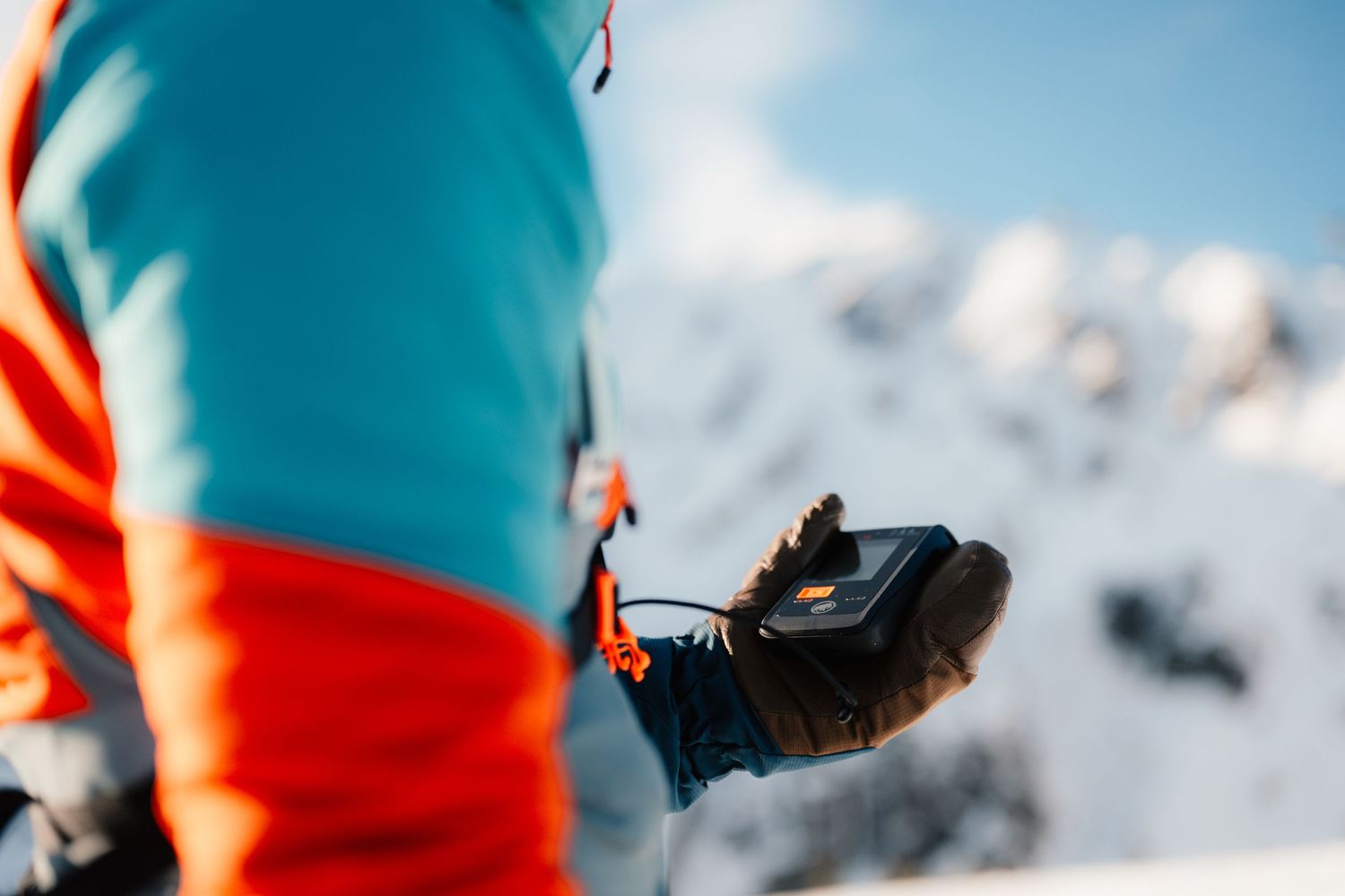 Close-up of skier in teal and orange jacket adjusting ski boot binding in snowy mountain terrain.