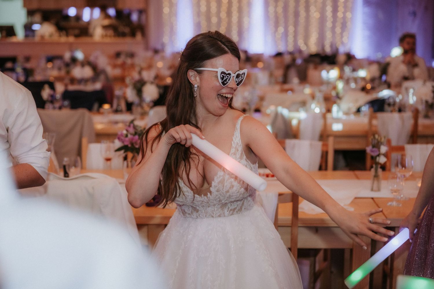 Bride in white gown and heart sunglasses dancing joyfully at elegant wedding reception venue.