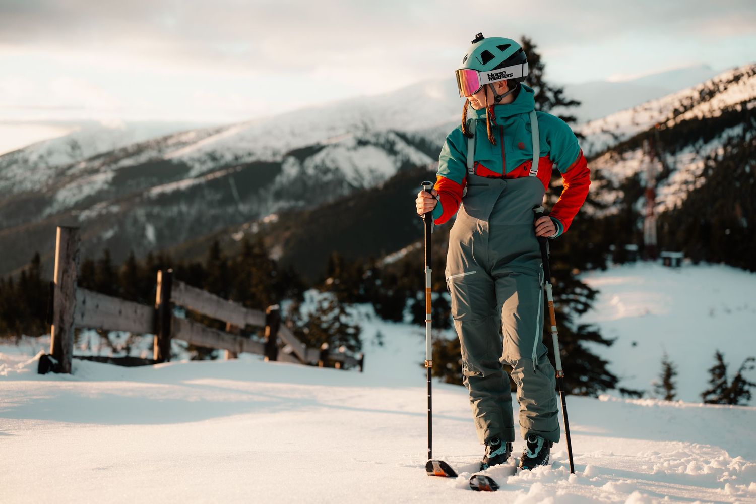 A skier in teal and red gear stands with poles on a snowy mountain slope at golden hour.