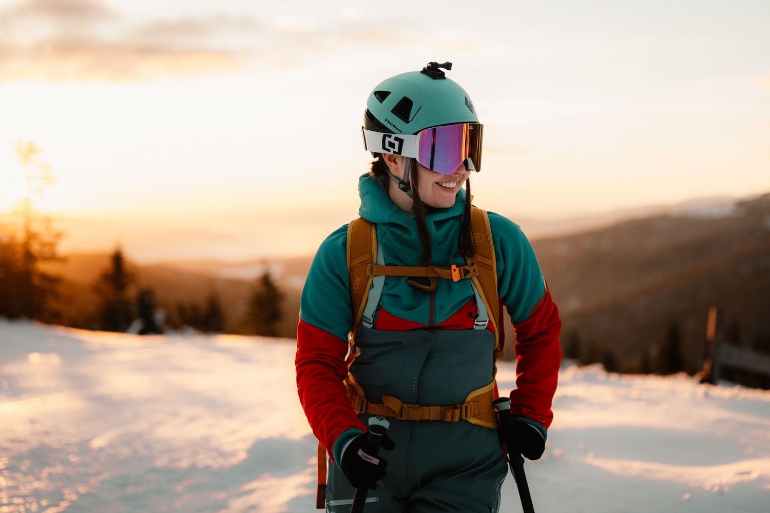 Smiling skier in teal helmet and goggles on snowy mountain slope at vibrant sunset.