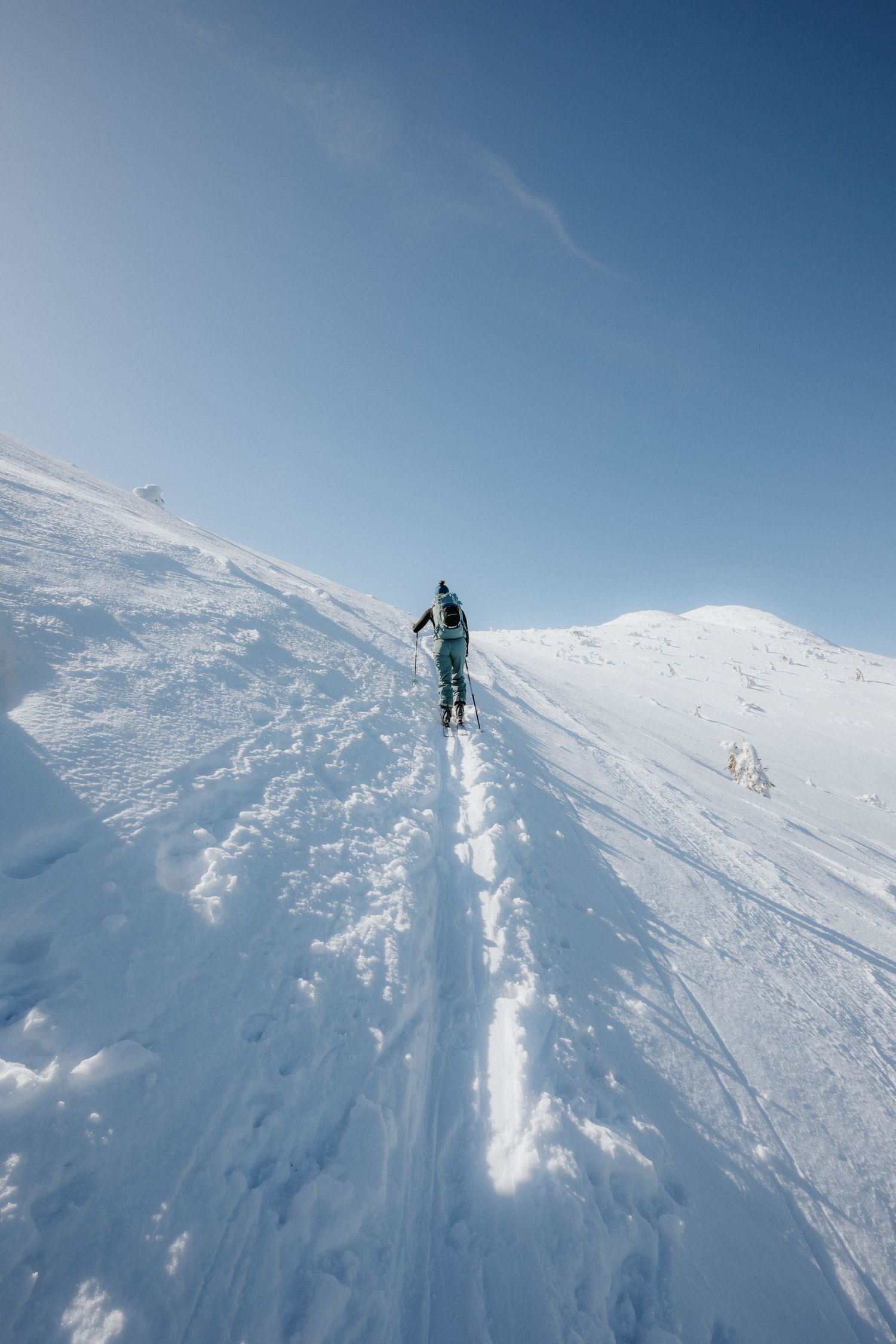 Lone backcountry skier climbing a vast open snowy mountain slope under a clear blue sky.