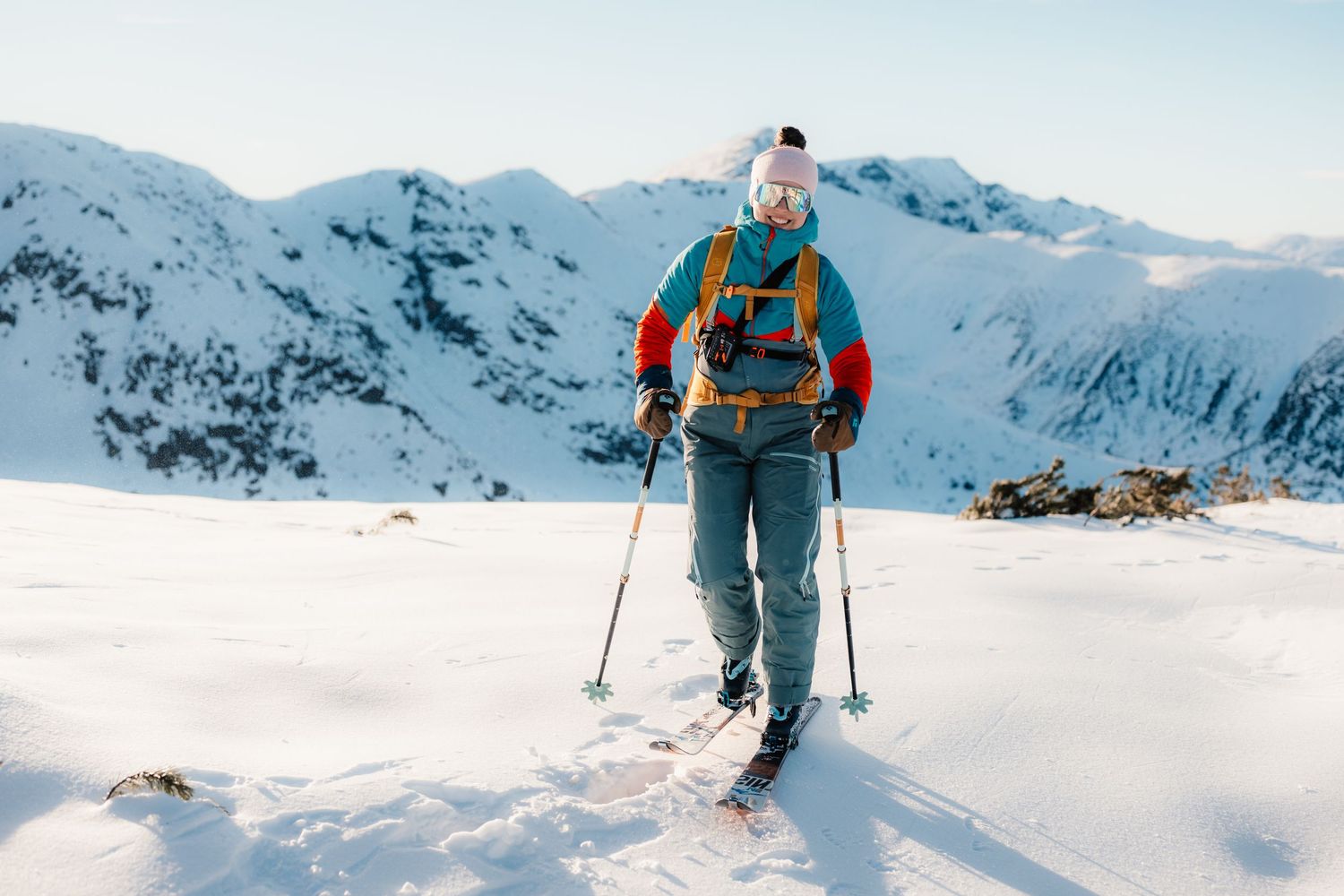 Backcountry skier with orange pack standing on vast snowy mountain terrain under a clear winter sky.