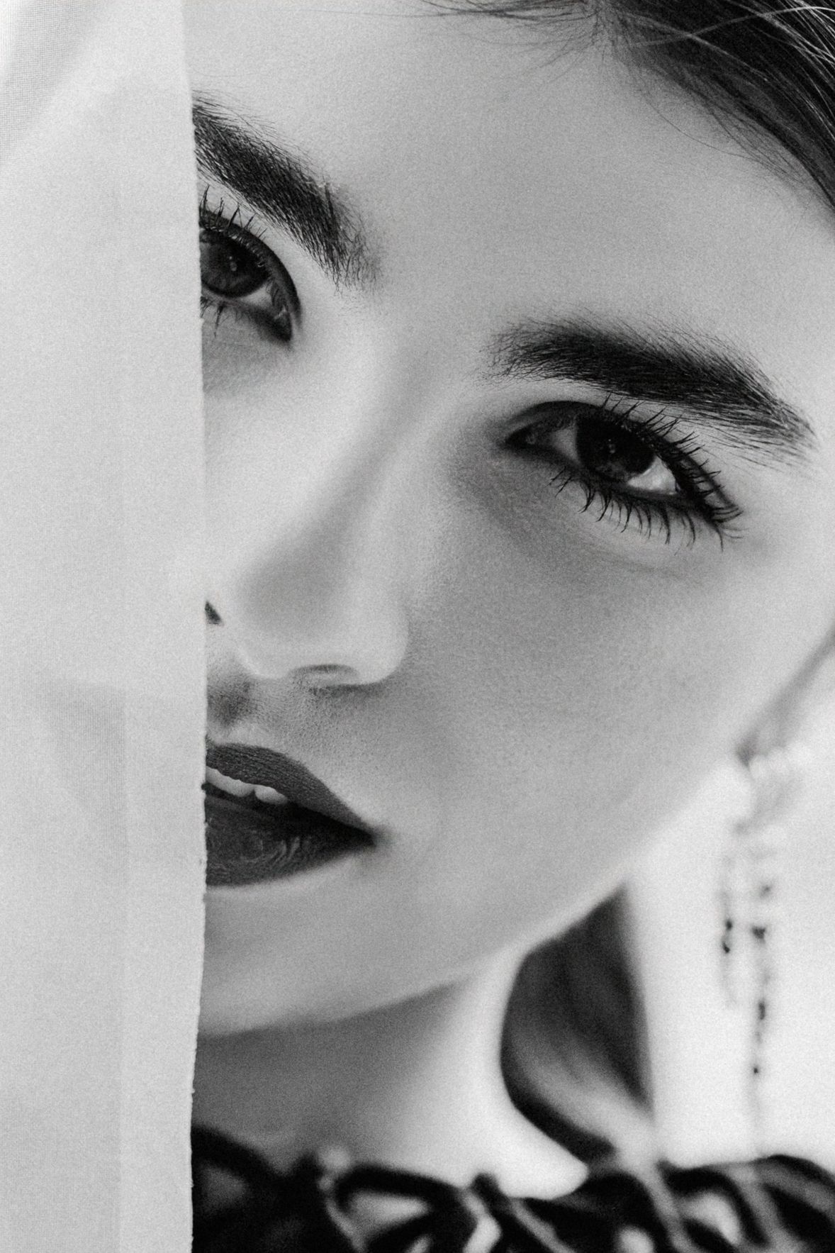 Black and white close-up portrait of a young woman with bold brows and dark lips, partially hidden behind sheer fabric.