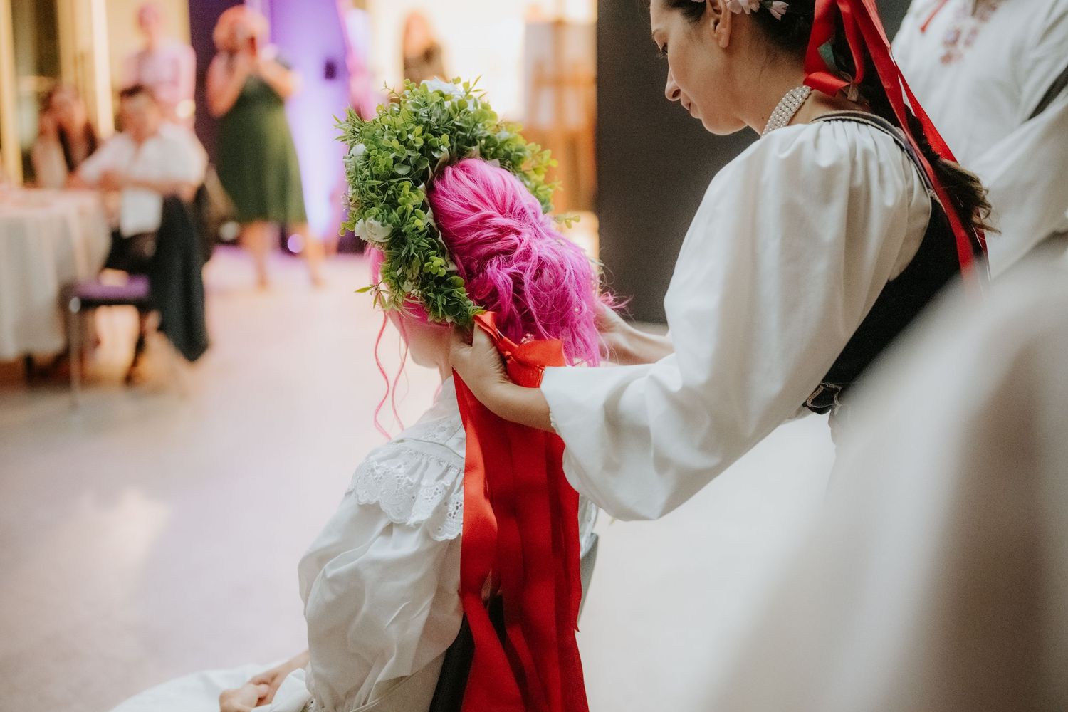 Woman placing a green floral wreath on a girl with pink hair in traditional folk costumes at a cultural event.