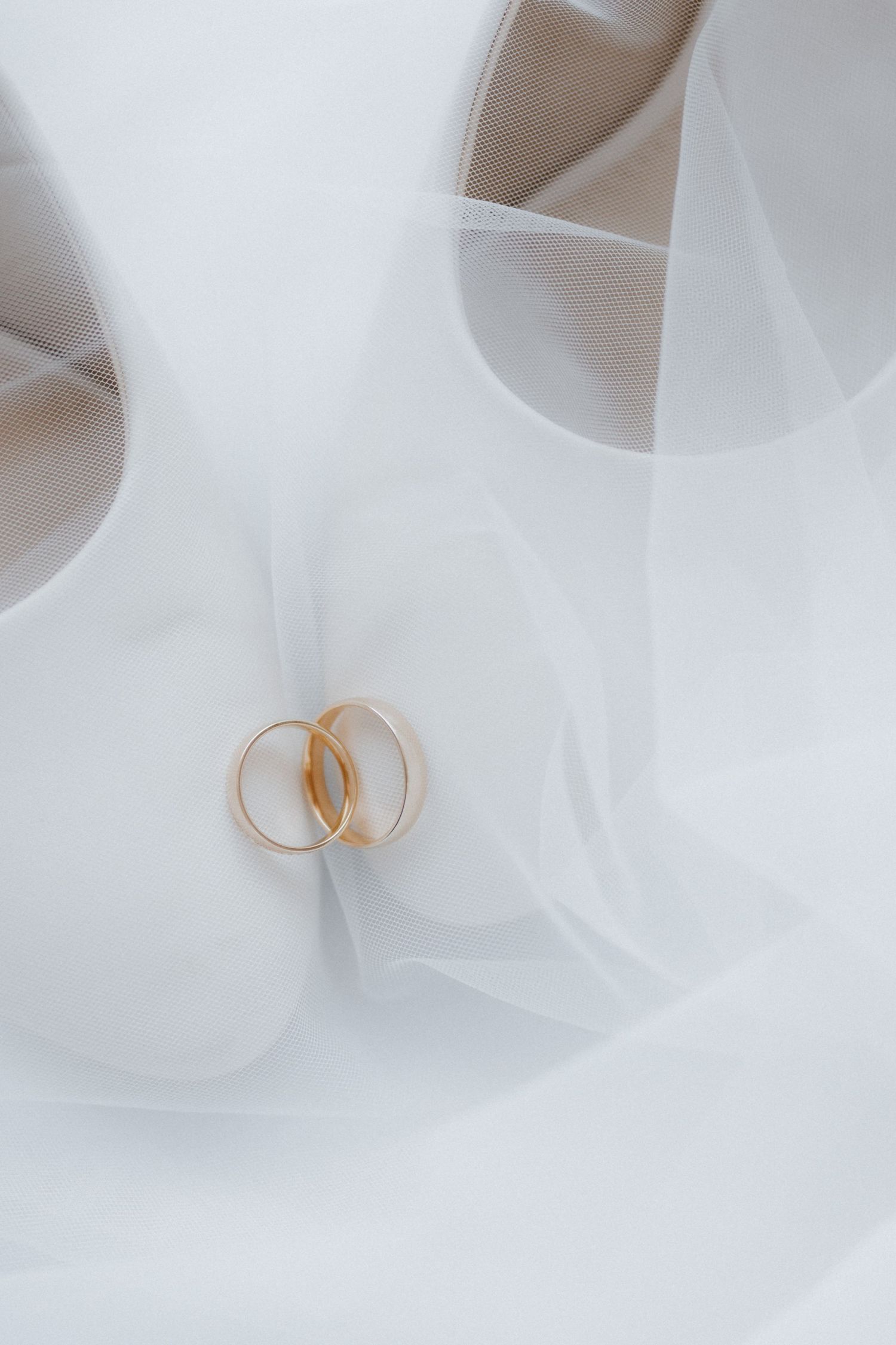 Two gold wedding bands resting on a white bridal veil beside elegant white heels.