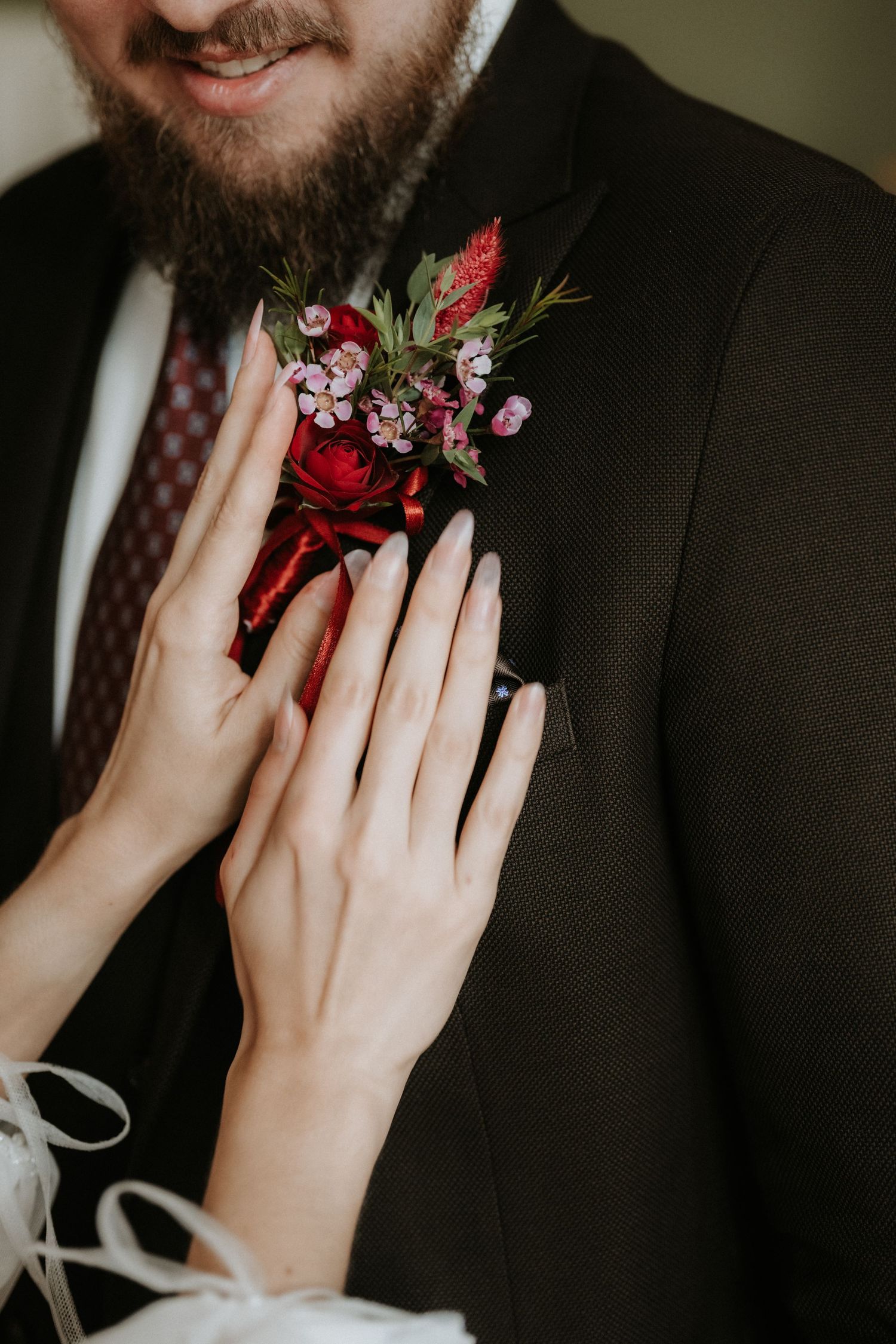 Bride adjusting a red rose boutonniere on groom's dark suit lapel, with delicate pink flowers and greenery.