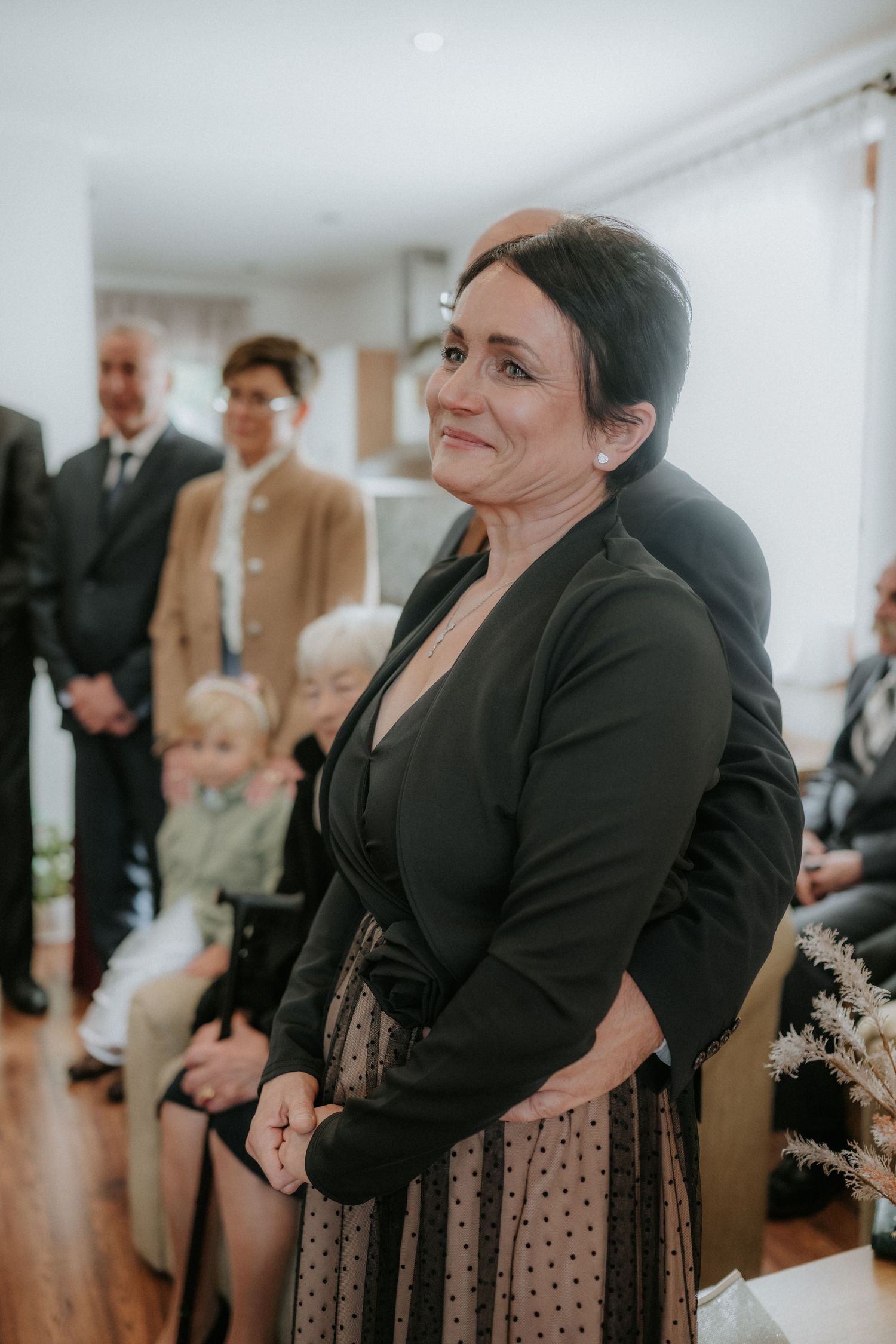 A woman in a black blazer and polka dot skirt smiles warmly at a formal indoor gathering.
