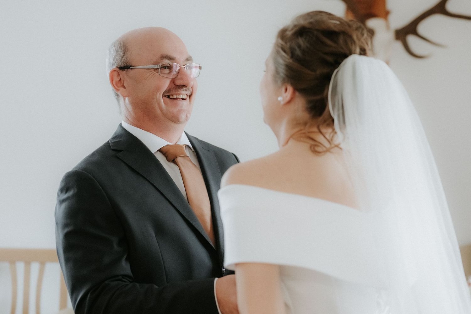 A smiling groom in a dark suit shares a tender first look moment with a bride in a white off-shoulder gown and veil.