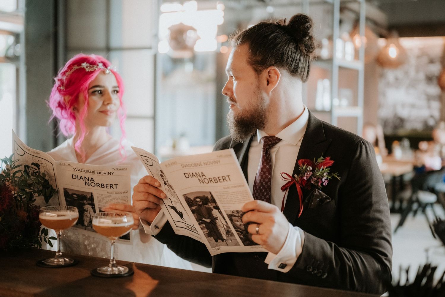 Bride with pink hair and groom in suit reading wedding newspapers together at a bar, with beer and floral boutonniere.