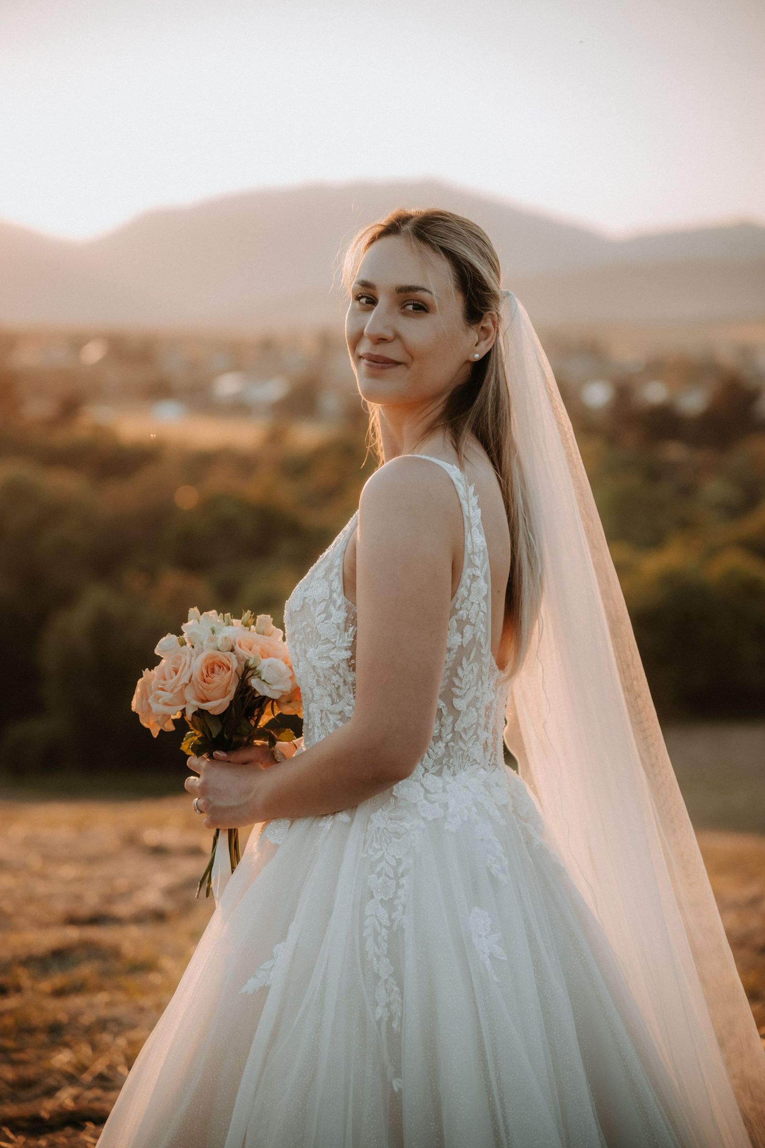 Bride in white lace ballgown and veil holds peach roses bouquet at golden hour with mountain backdrop.