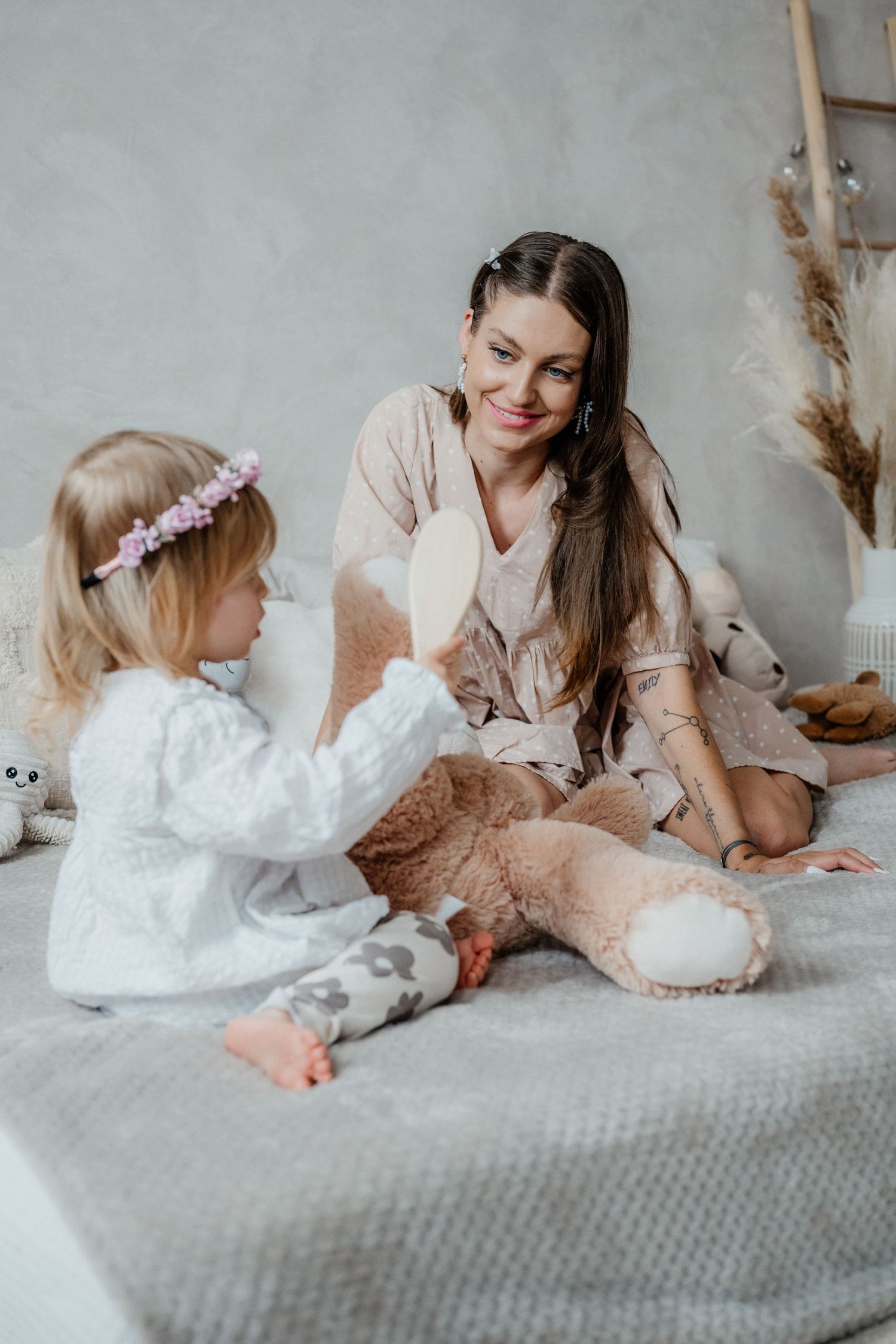 Smiling mother sitting on bed watching toddler daughter hold a wooden hairbrush.