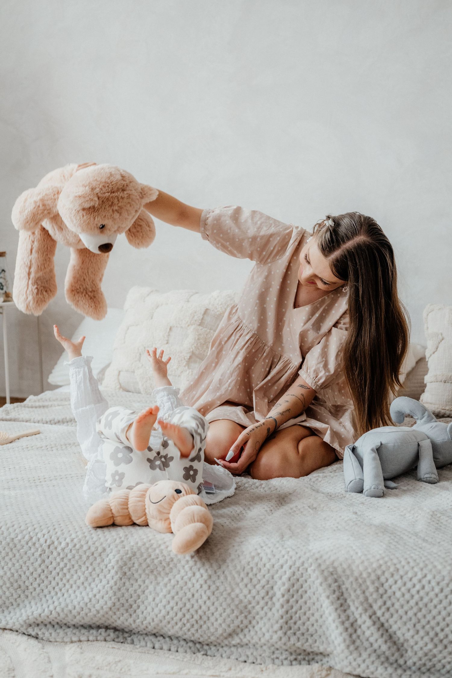 Brunette mother sitting on bed holding large teddy bear over smiling baby in soft nursery setting.