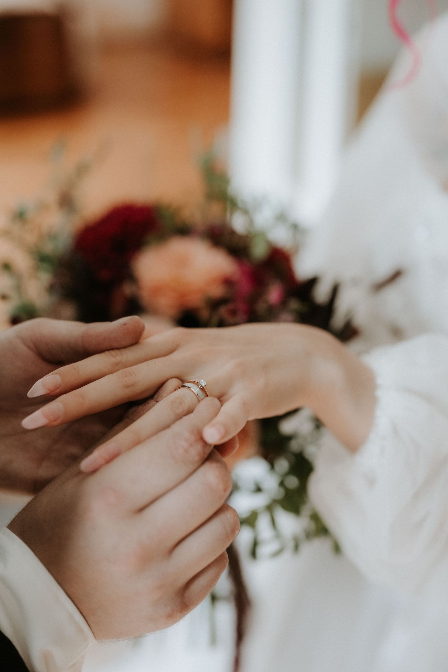 Close-up of a couple exchanging wedding rings with a bridal bouquet of red and burgundy flowers in the background.