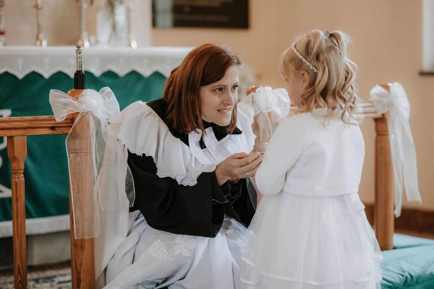 Woman in black dress kneeling beside young girl in white dress inside a church during First Communion.