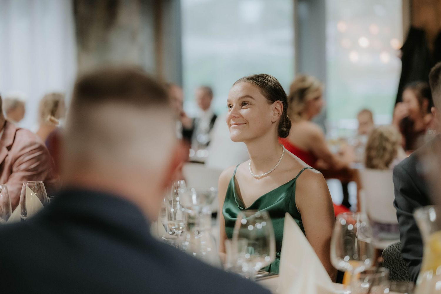 Woman in green dress smiling at wedding reception dinner, wearing pearl necklace, seated at elegantly set table.