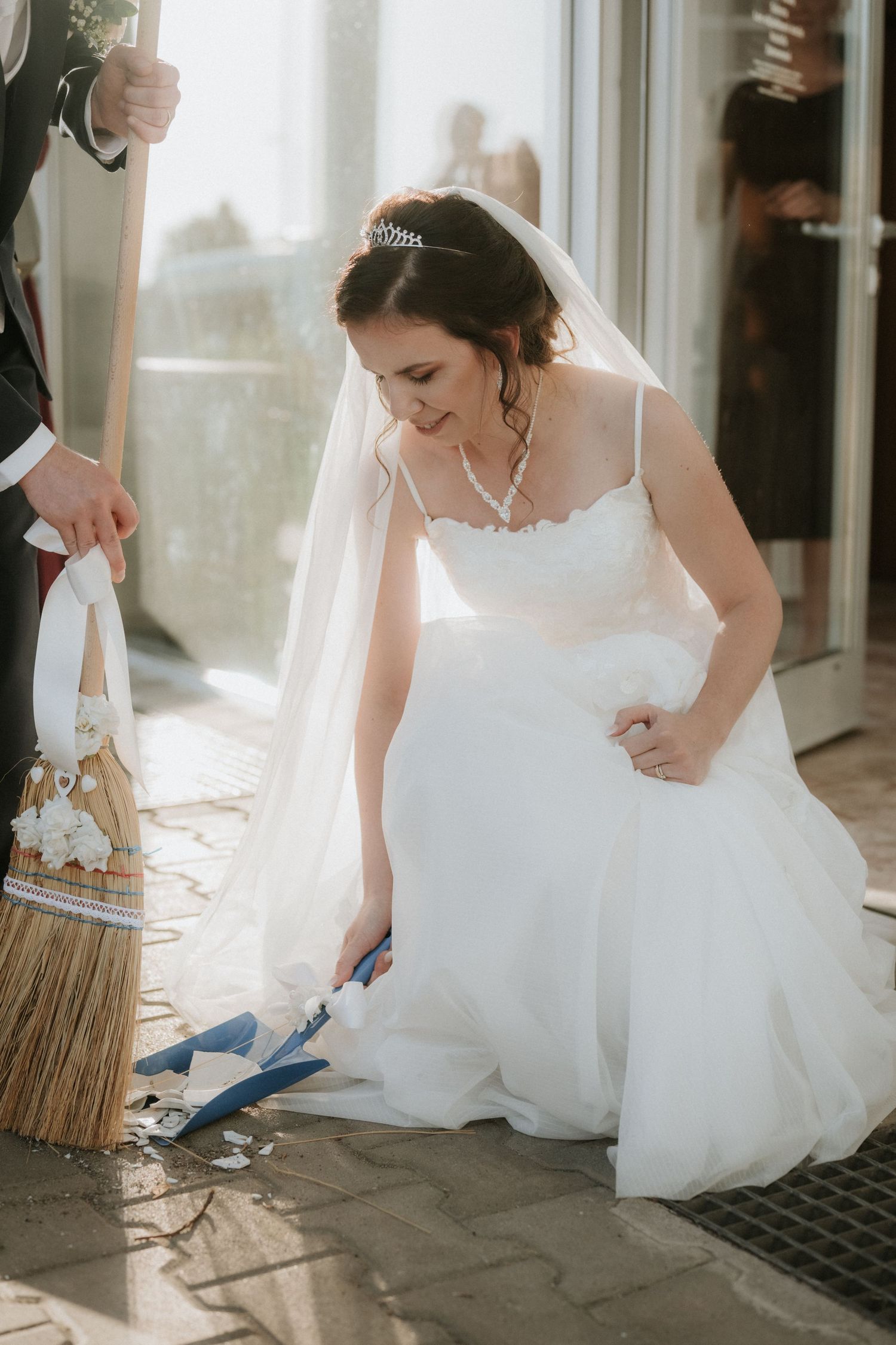 Bride in white gown kneeling to sweep broken glass with a broom during a wedding tradition ceremony.