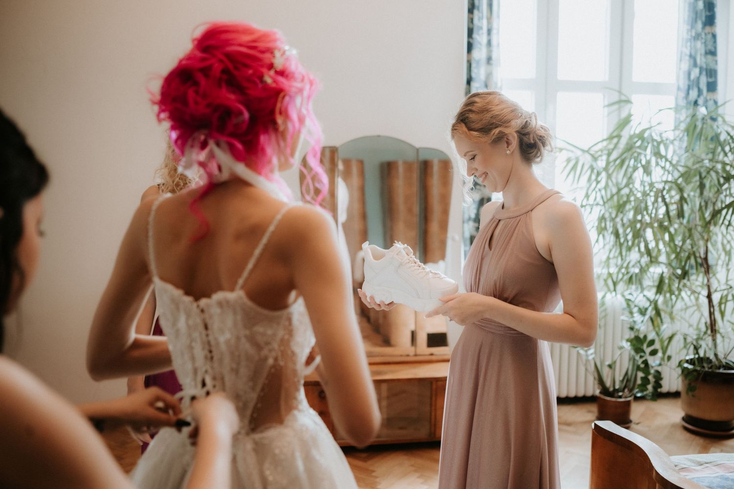 Bride with pink hair in a lace wedding dress being helped by a bridesmaid in a mauve gown in a bright room.