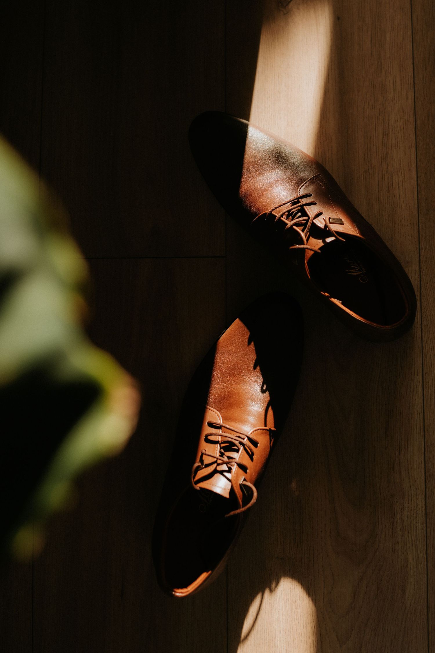 A pair of tan leather Oxford dress shoes with laces resting on a wooden floor in warm sunlight.