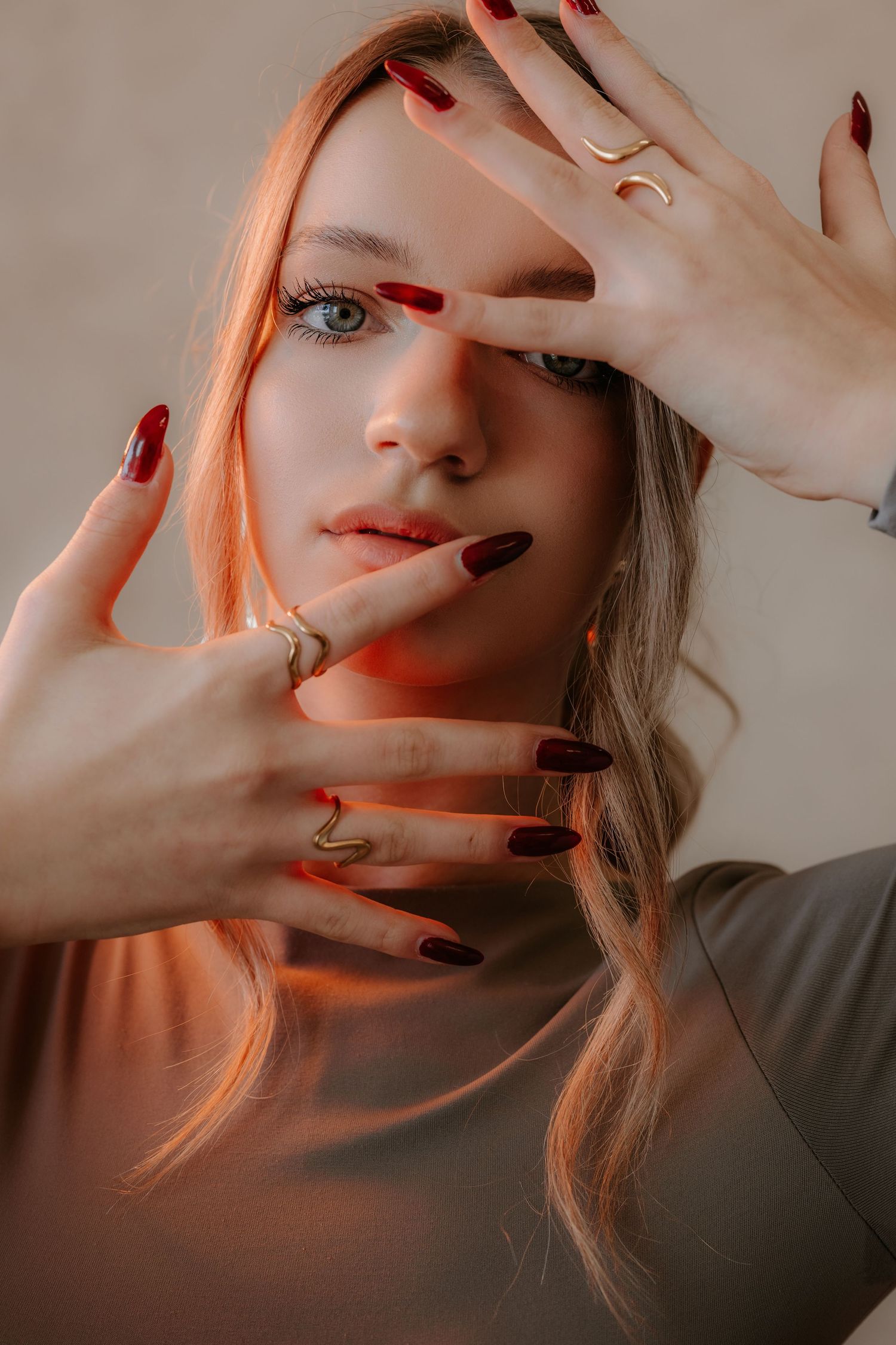 Close-up portrait of blonde woman with dark red nails and gold rings framing her face in warm light.