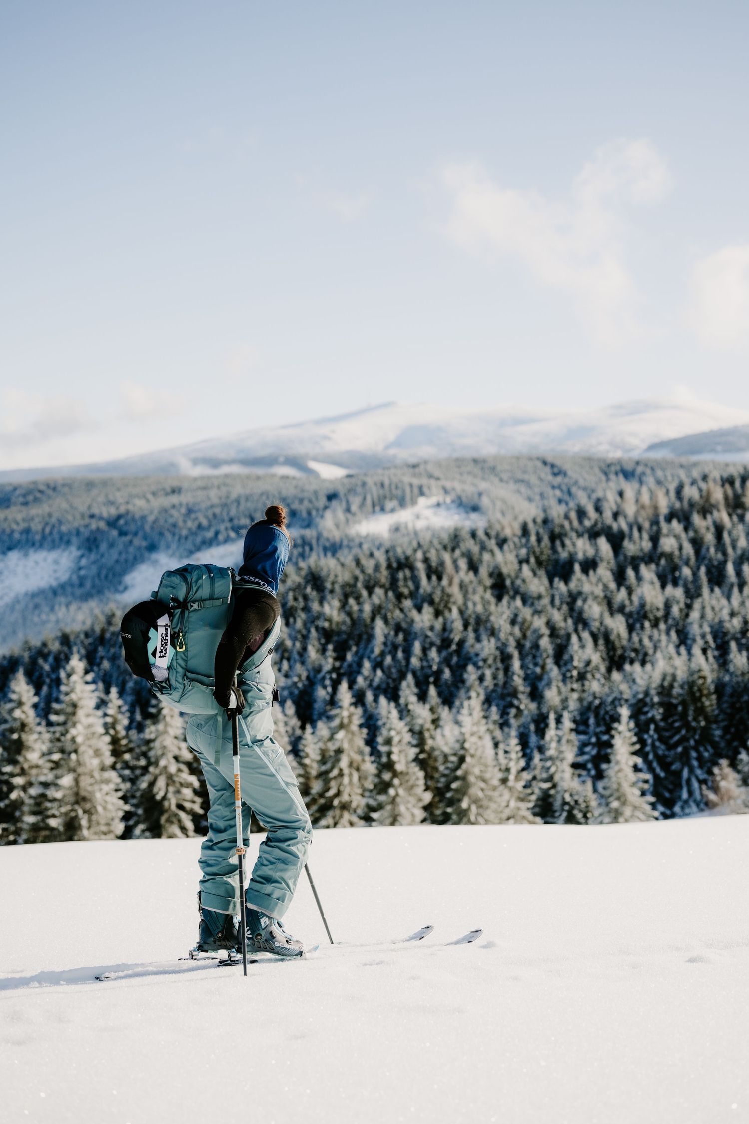 A backcountry skier pauses on a snowy hillside overlooking a vast snow-covered forest and mountain range.