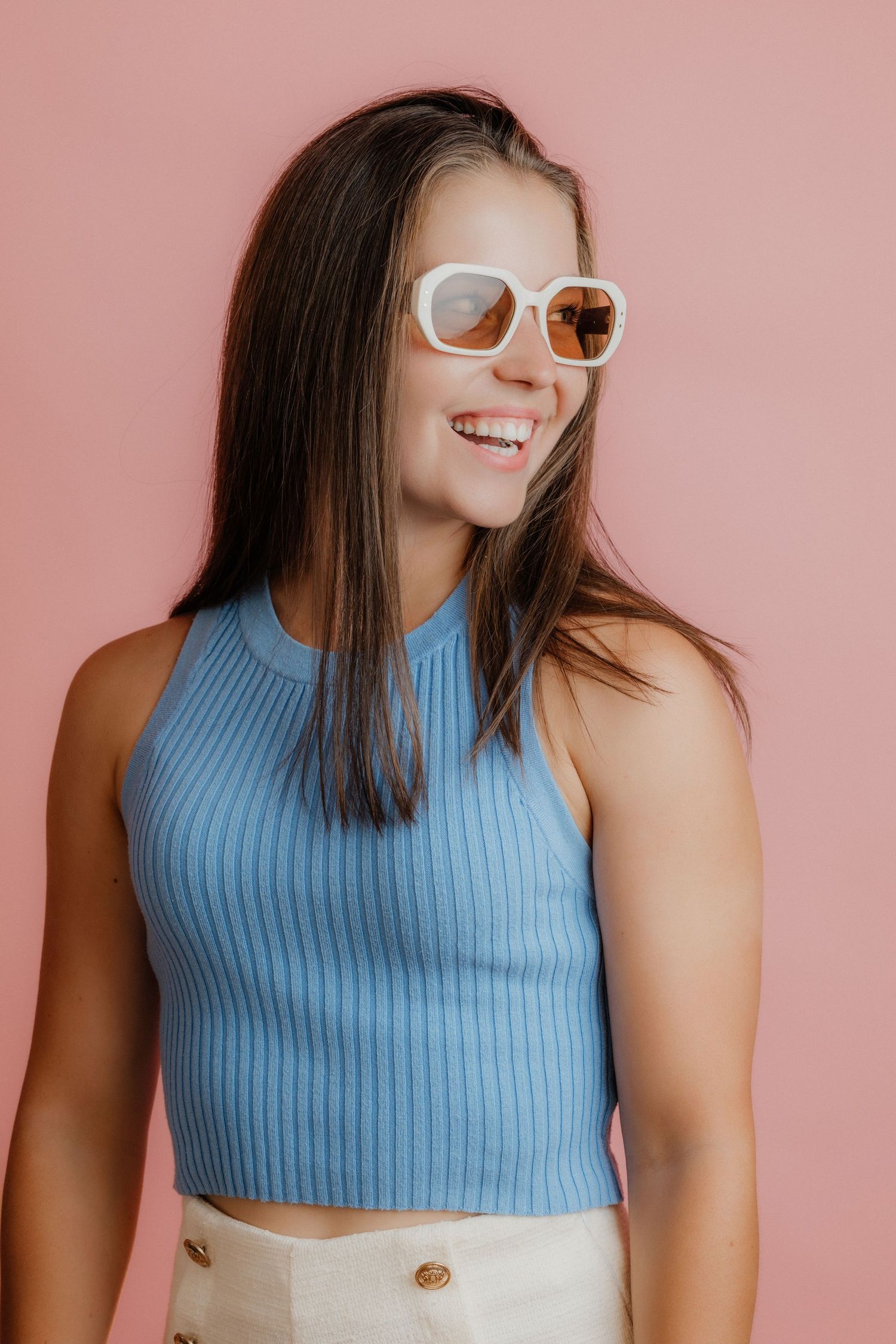 Smiling young woman wearing white sunglasses and a blue ribbed crop top against a pink background.