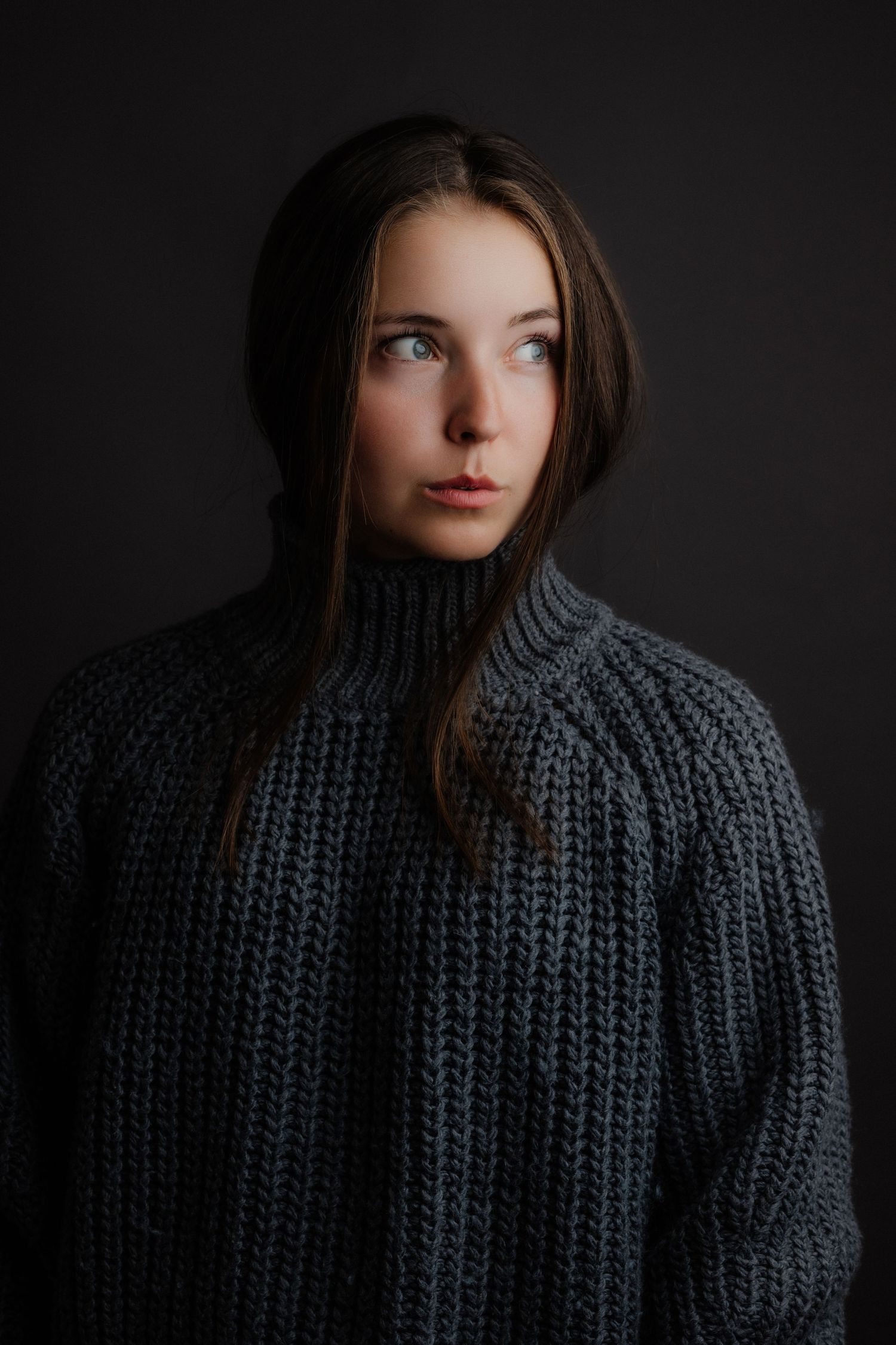 Young woman in an oversized chunky grey turtleneck sweater looking upward against a dark studio background.