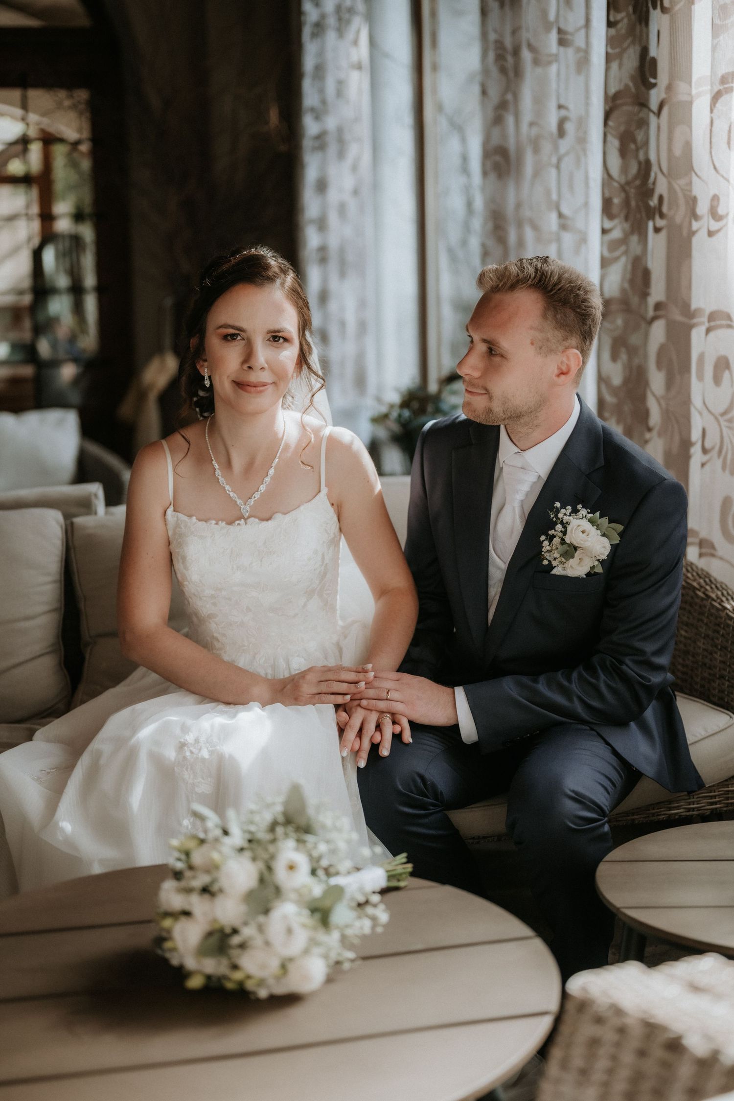 Newlyweds sitting on a couch holding hands with a white floral bouquet on the table nearby.